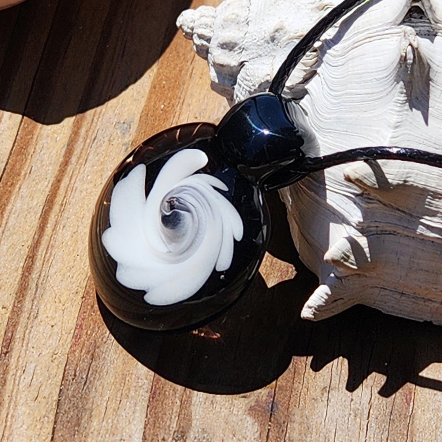 Artistic black and white swirl glass pendant necklace placed on wood with shell, showcasing its handmade craftsmanship and bold contrast design.