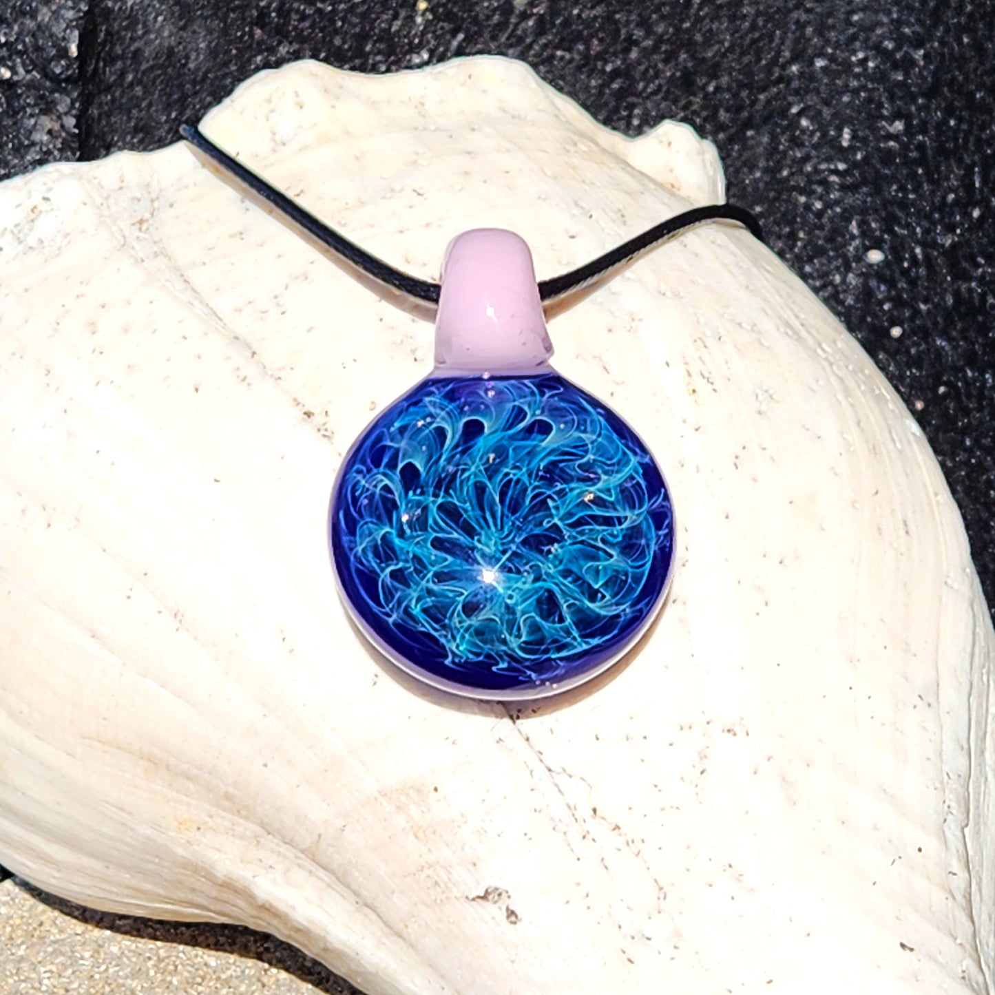 Handmade pendant necklace with intricate blue glass art and pink loop, displayed on a seashell. Unique blown glass charm for jewelry lovers.