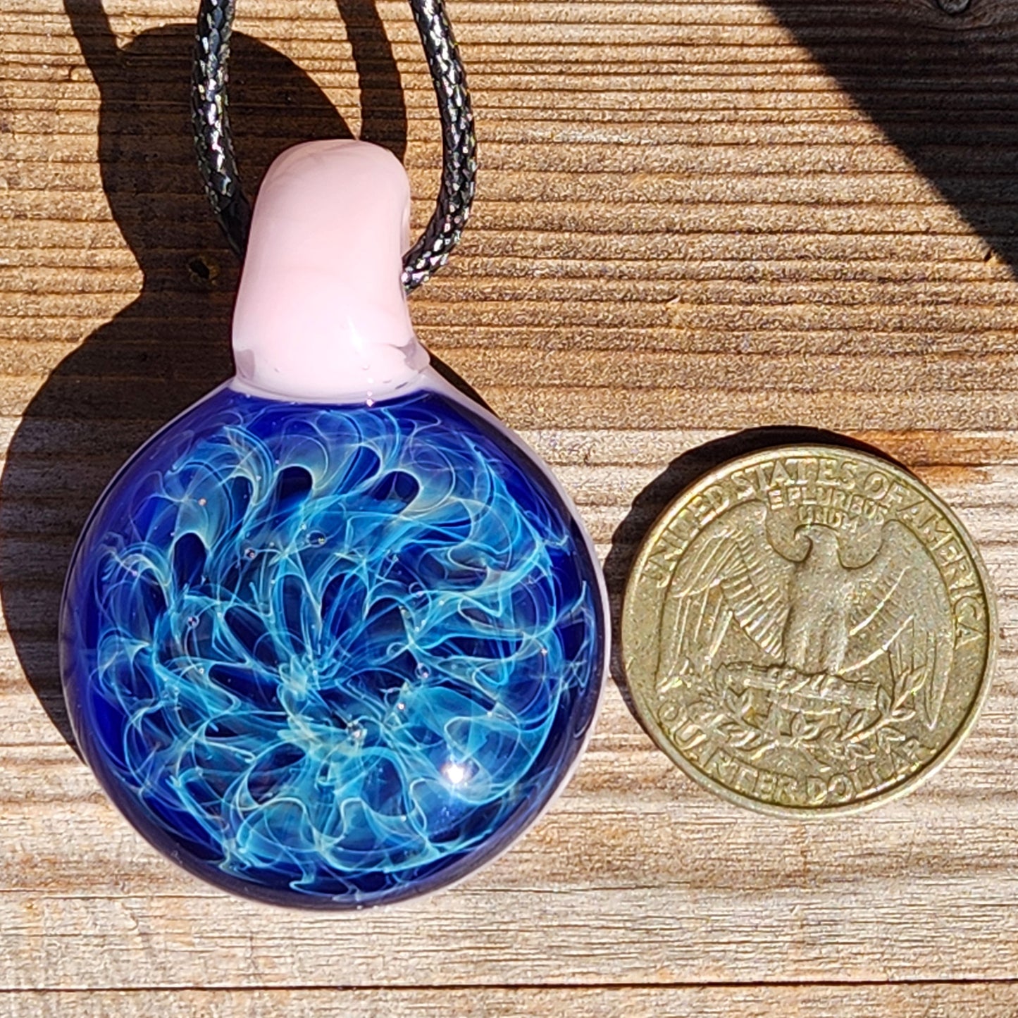 Size comparison of handmade glass pendant necklace next to a US quarter. Heady pendant with pink loop and blue fumed design.