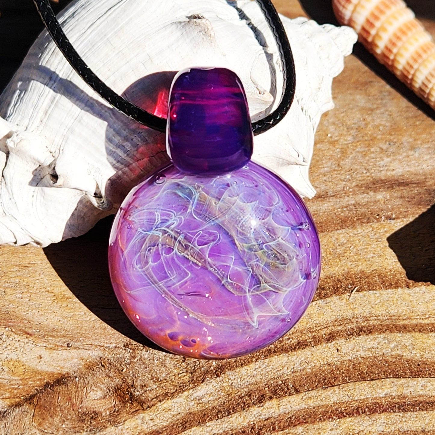 Purple glass pendant necklace with silver and gold fuming, displayed on wood with seashells. Bold artisan jewelry in vivid purple tones.