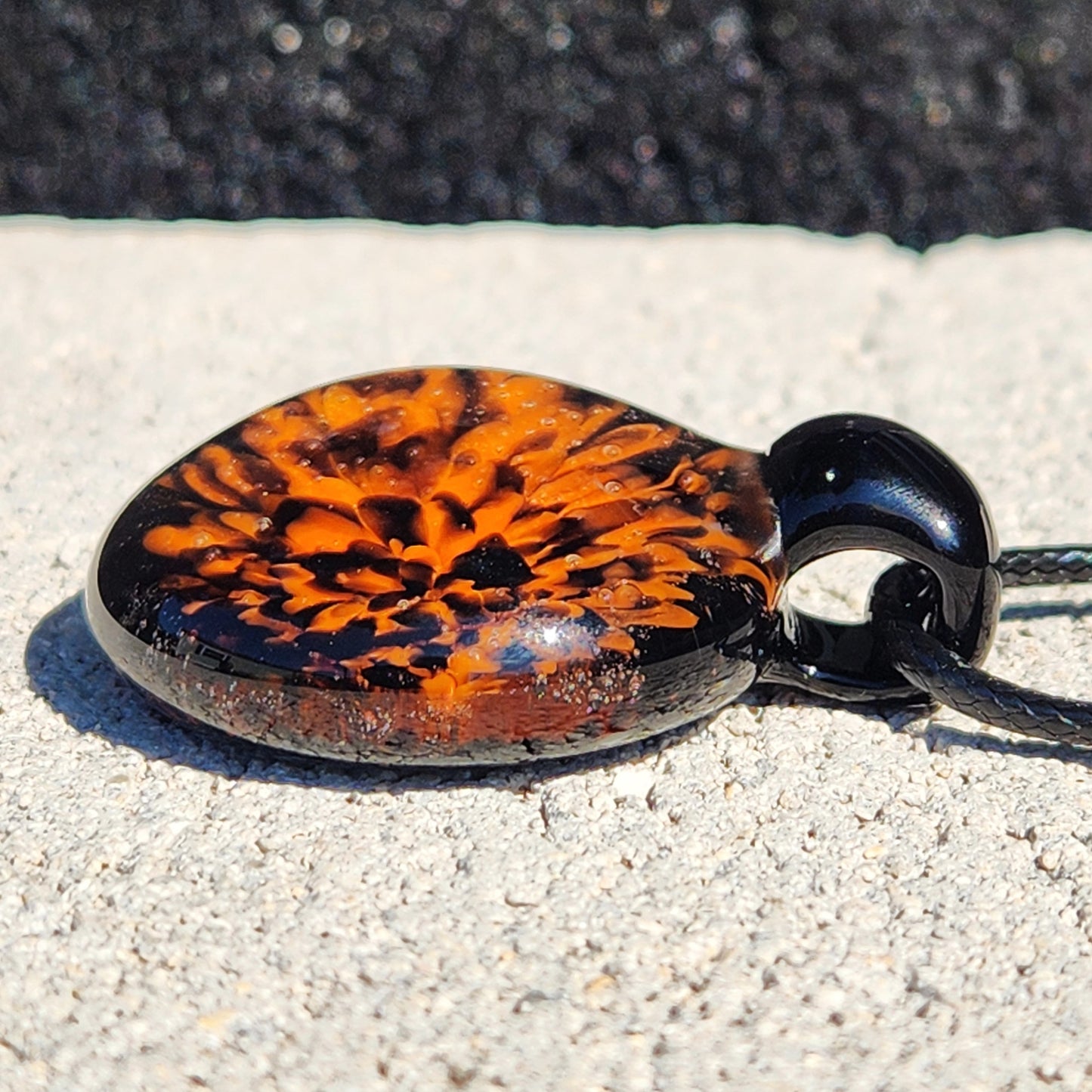 Fiery Glass Pendant Necklace lying on stone, showcasing the detailed orange fire bloom and glossy black finish. Perfect handmade Halloween accessory.