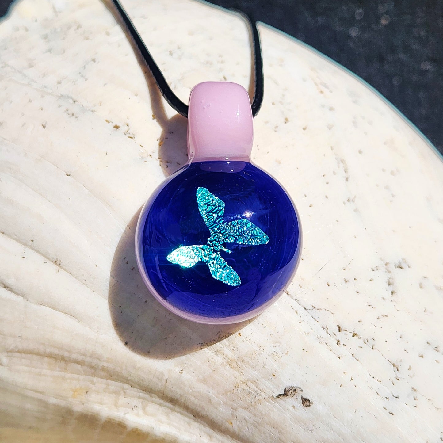 Small glass butterfly pendant with shimmering blue dichroic design and pink loop, displayed on a seashell. Handmade jewelry gift.
