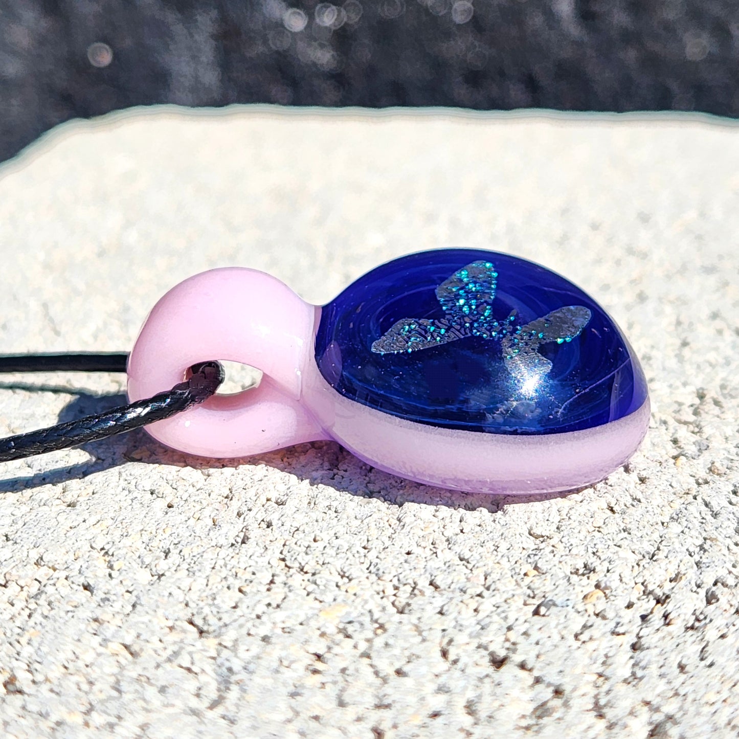 Close-up of handmade small glass pendant featuring a sparkling blue butterfly and soft pink loop, lying on textured stone.