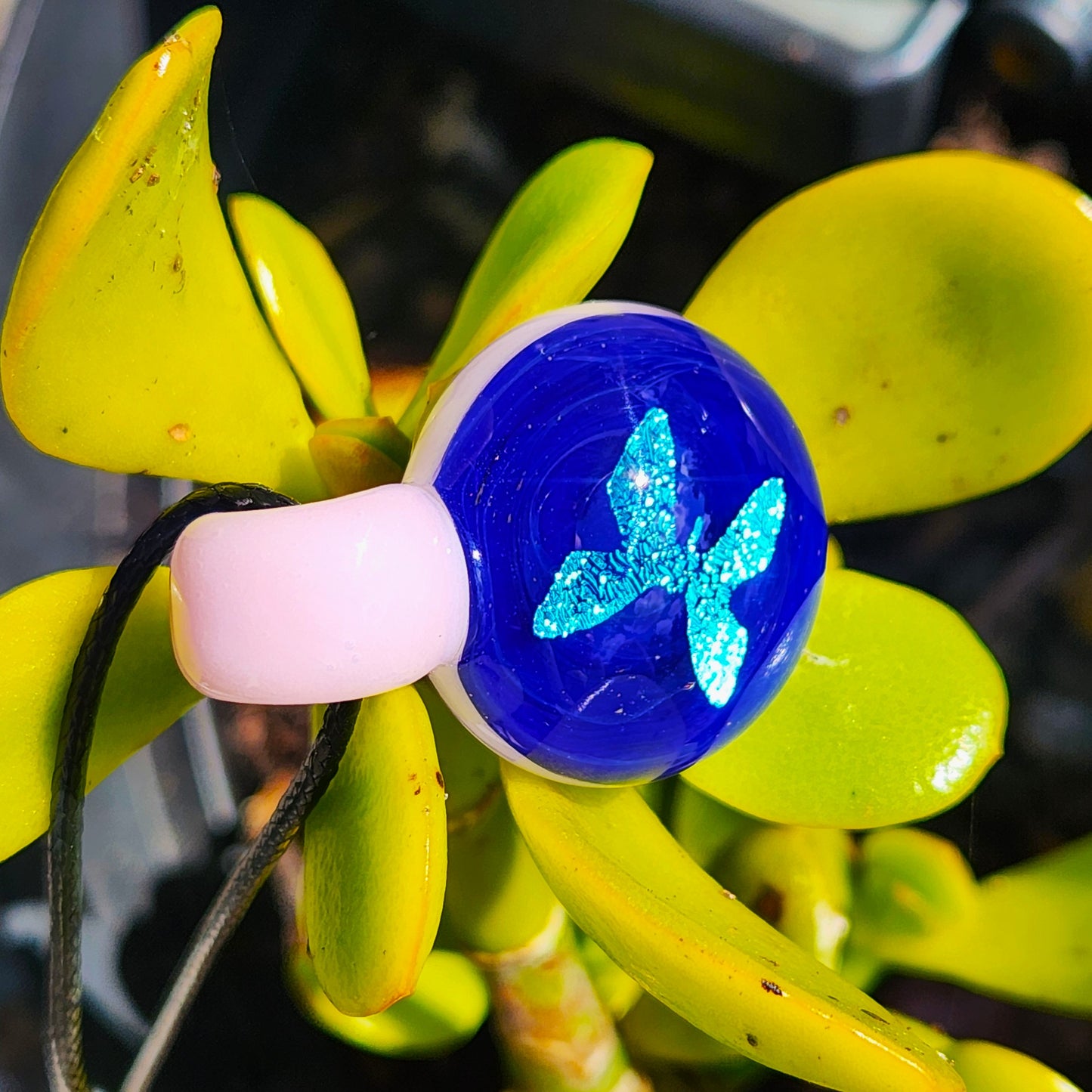 Blue butterfly glass pendant with pink loop resting on plant leaves, showcasing its sparkle and handmade charm.