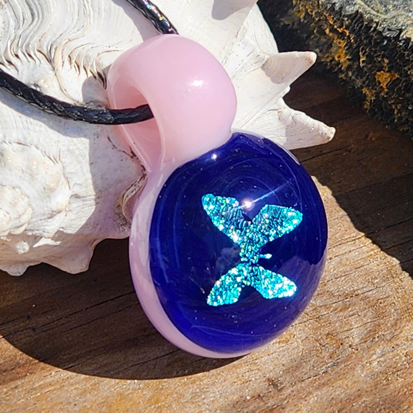 Handmade blue butterfly glass pendant with pink loop, displayed near a seashell on rustic wood for natural jewelry presentation.