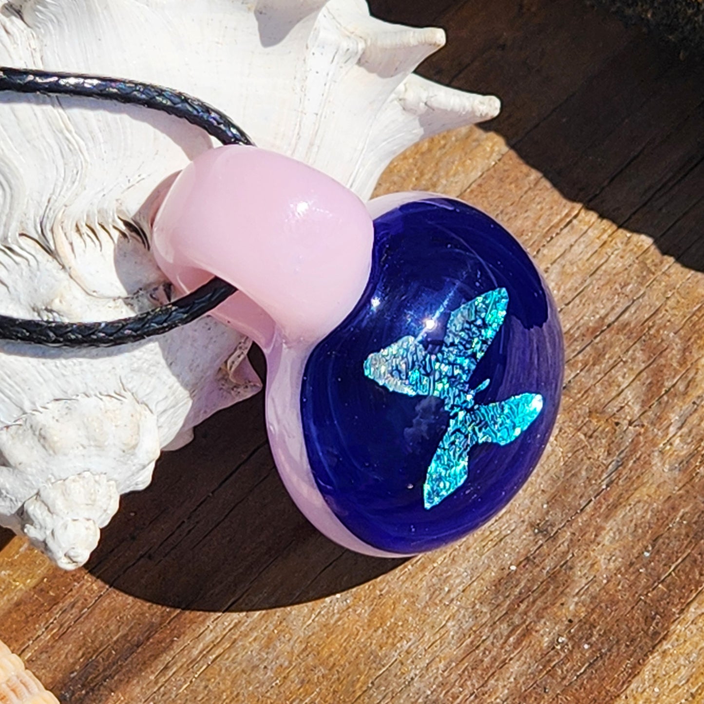 Blue dichroic butterfly pendant with pink glass loop on black cord, resting on a wooden surface with seashell accent.