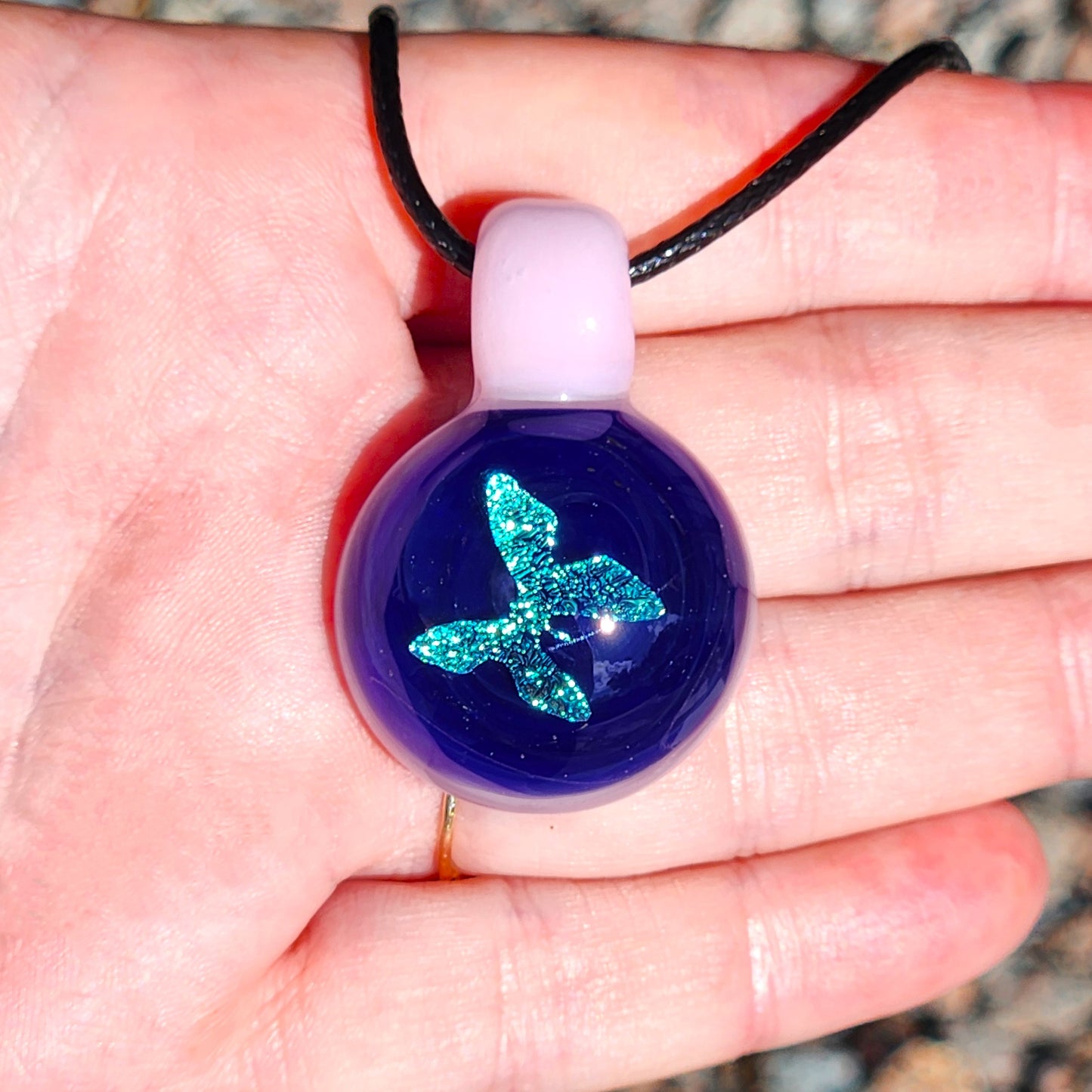 Hand holding small glass butterfly pendant with shimmering blue design and pink loop, highlighting its delicate size and detail.