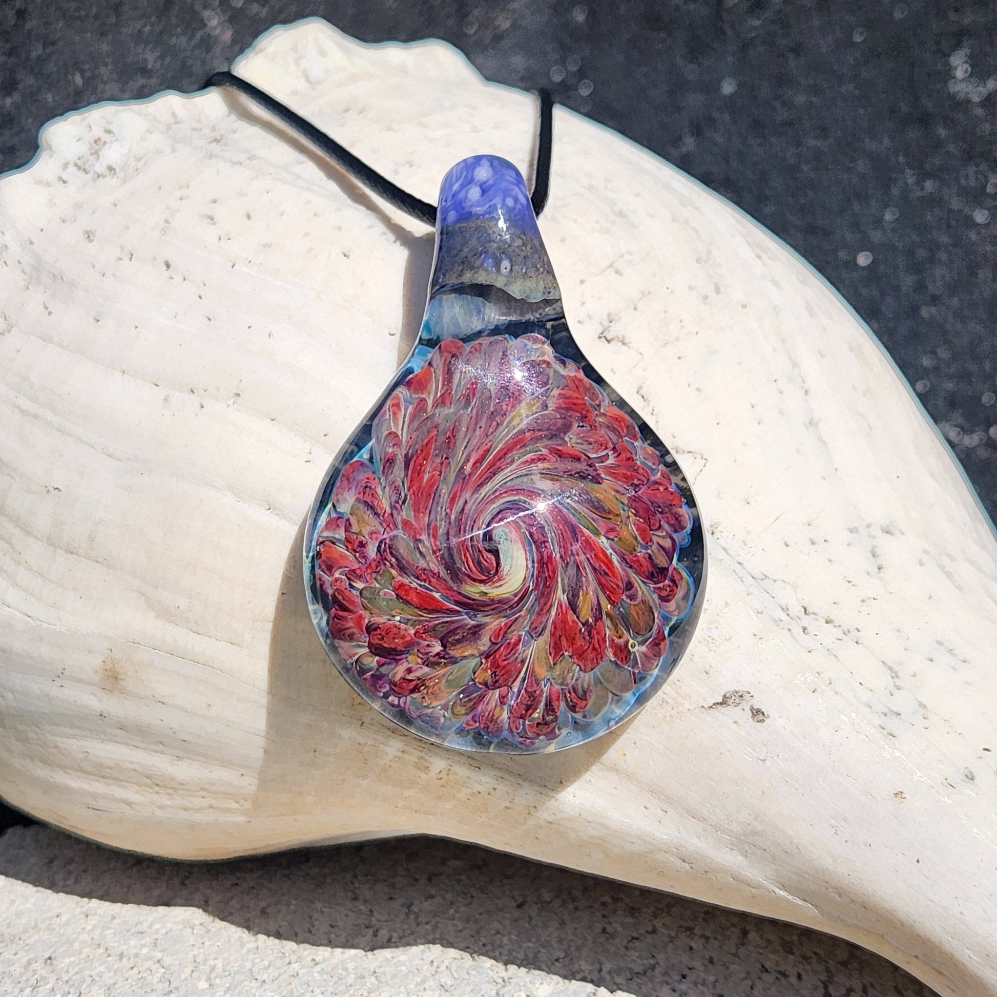Handmade heady glass pendant necklace with swirling red and amber design, displayed on a seashell. Unique handblown borosilicate glass jewelry.