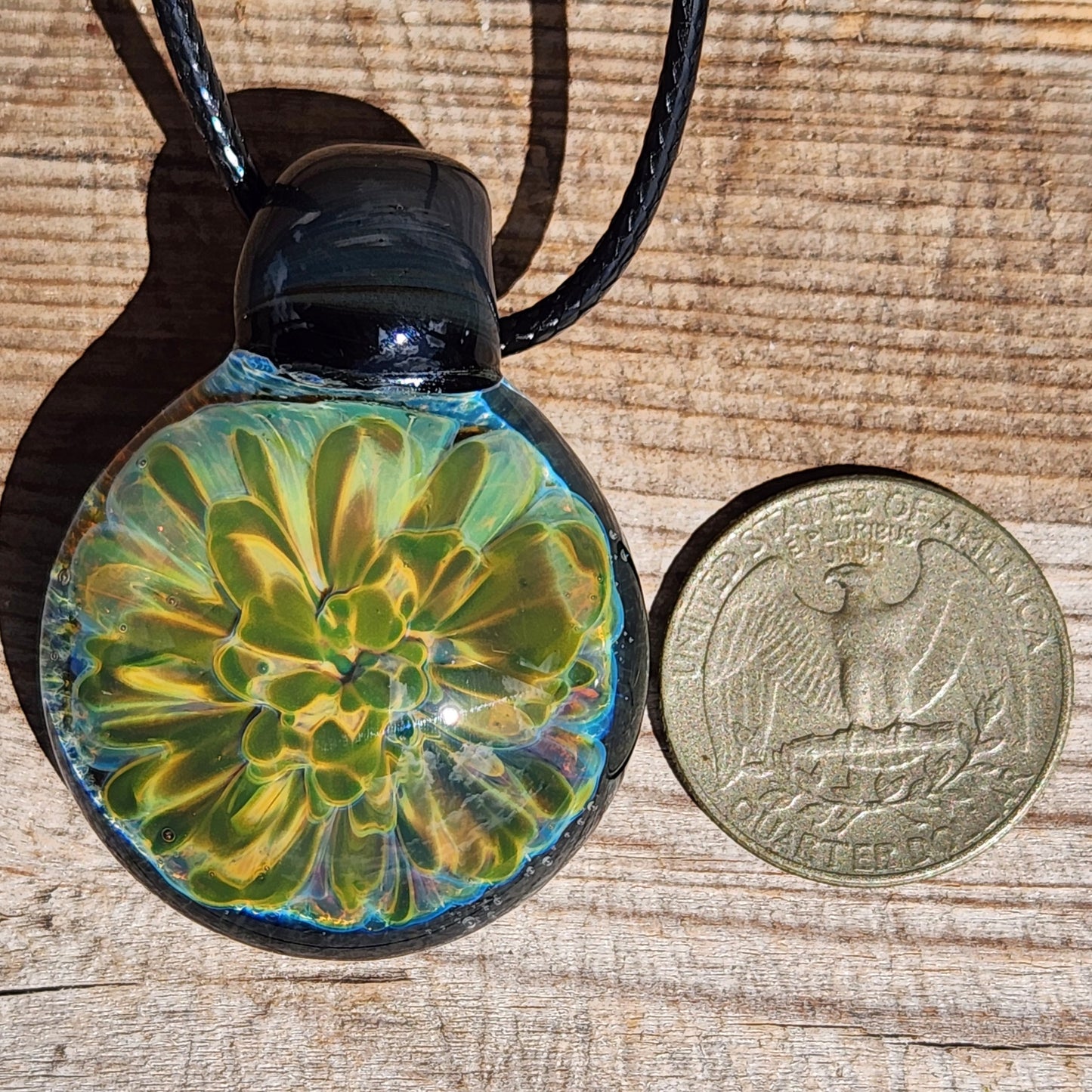 Handmade glass pendant with yellow-green flower next to a US quarter for size comparison. Unique necklace in gift box.