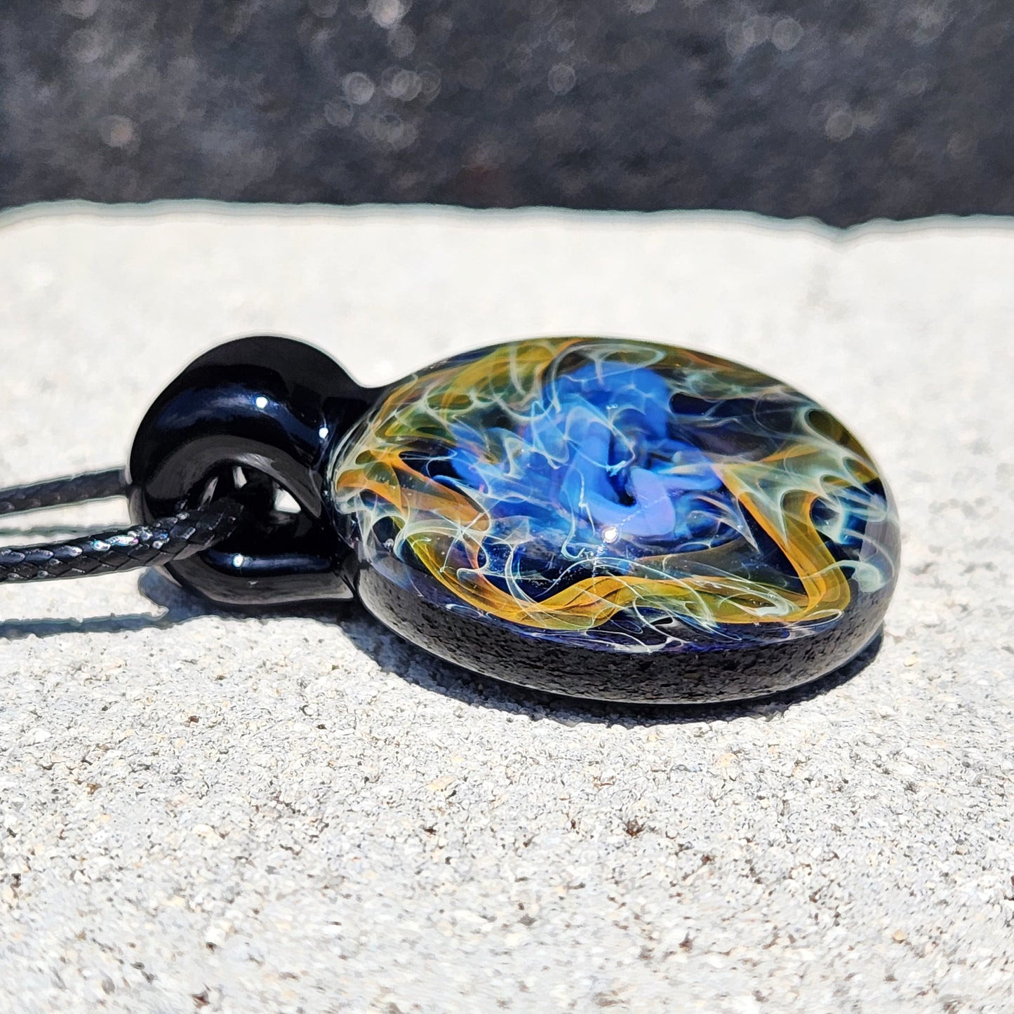 Close-up of gold and silver fumed glass pendant lying flat, showcasing intricate cosmic swirl design in blue, yellow, and white hues.