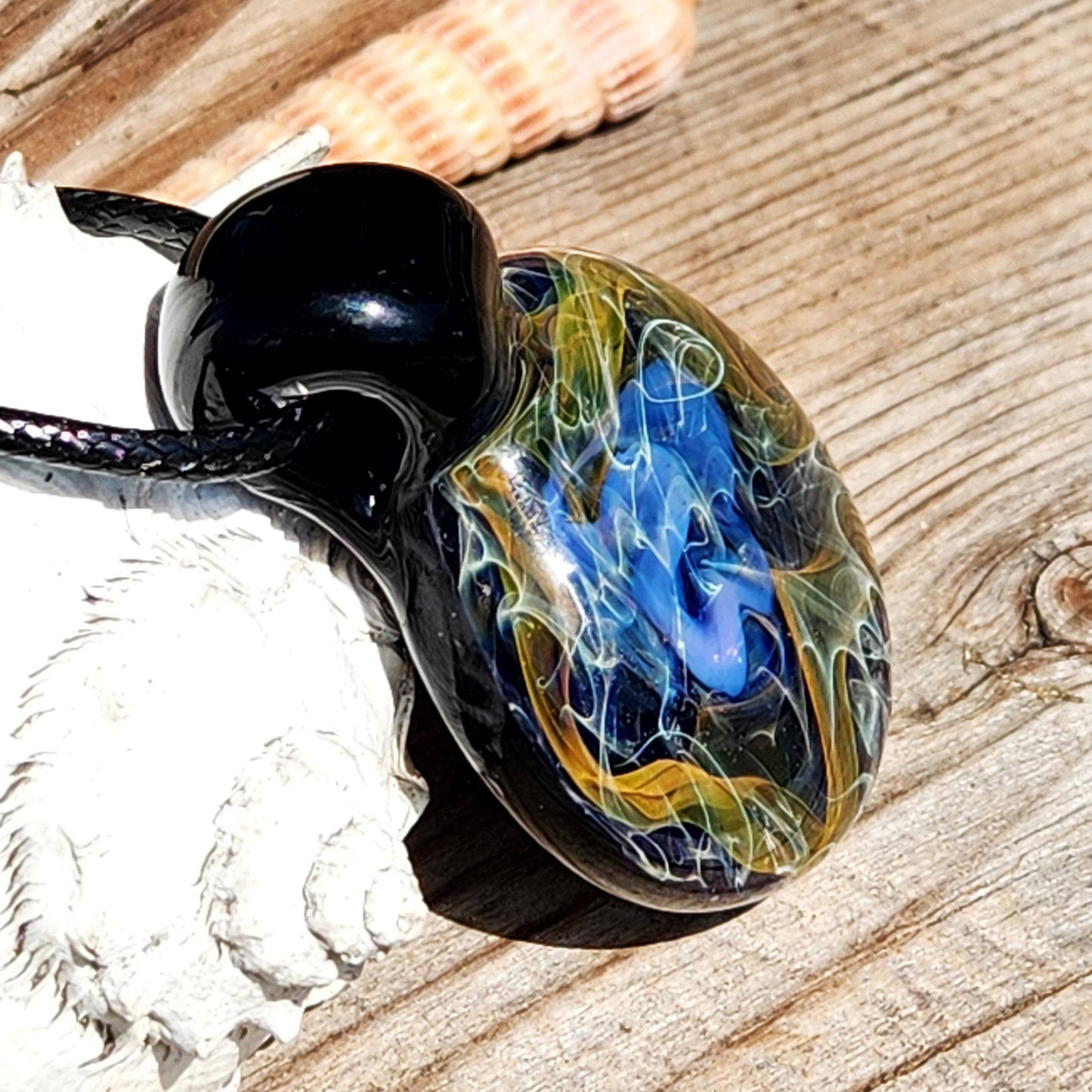 Handmade gold and silver fumed glass pendant resting on wood, featuring swirling cosmic blues and yellows for a unique jewelry accent.