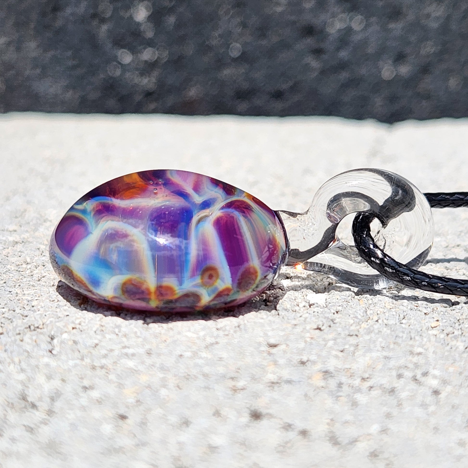Side profile of unique handmade borosilicate pendant with swirling rainbow hues, attached to black cord for wearable glass art.