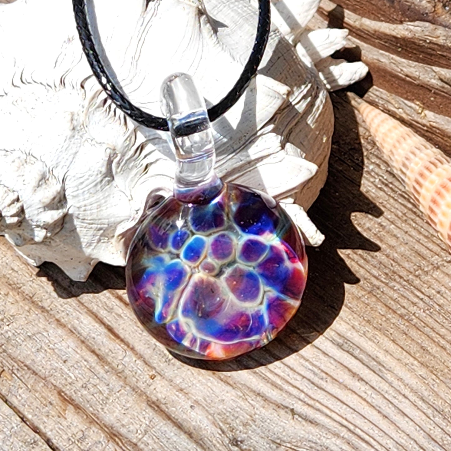 Colorful handmade glass pendant with blue, purple, and red hues resting on seashell, ideal for beachy boho jewelry lovers.