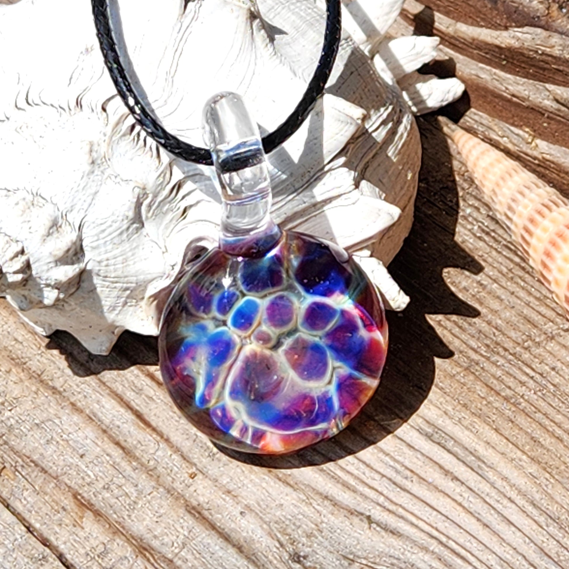 Colorful handmade glass pendant with blue, purple, and red hues resting on seashell, ideal for beachy boho jewelry lovers.