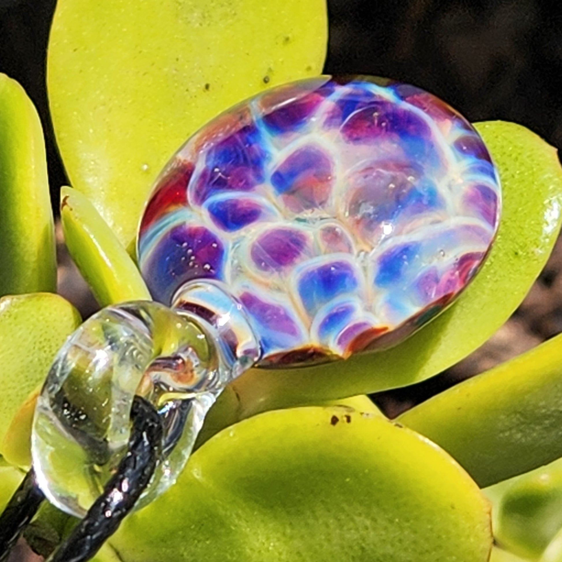 Vibrant purple and blue handmade pendant with bubble dot design, lying on succulent leaves, showcasing artistic borosilicate craftsmanship.
