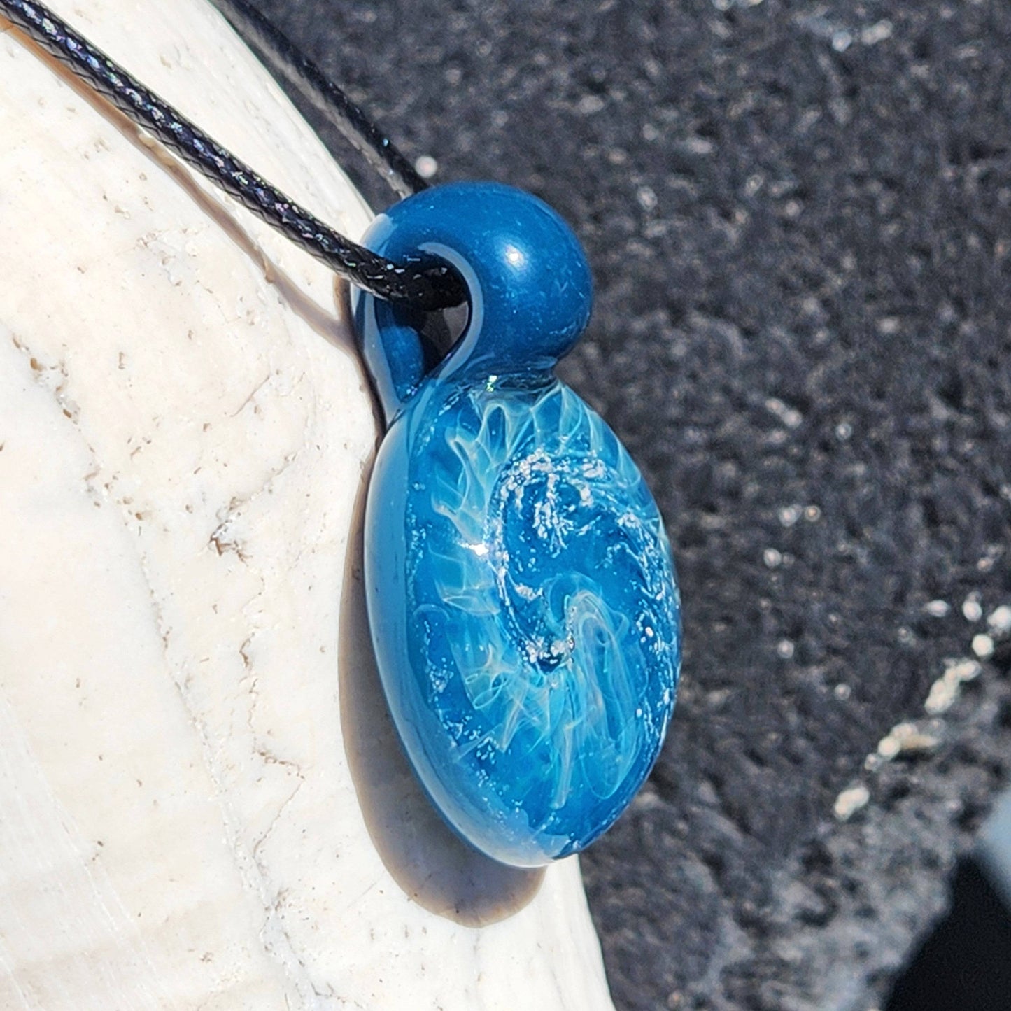 Ocean-inspired handmade glass pendant with swirling blues and silver, hanging on a black cord against a shell background.
