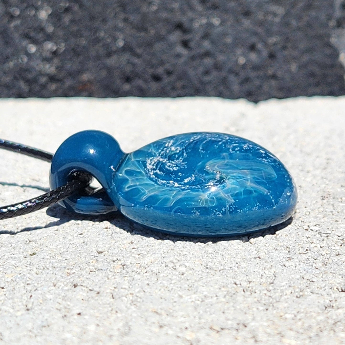 Side view of ocean-inspired glass pendant showing depth and silver shimmer, lying on a textured stone surface.