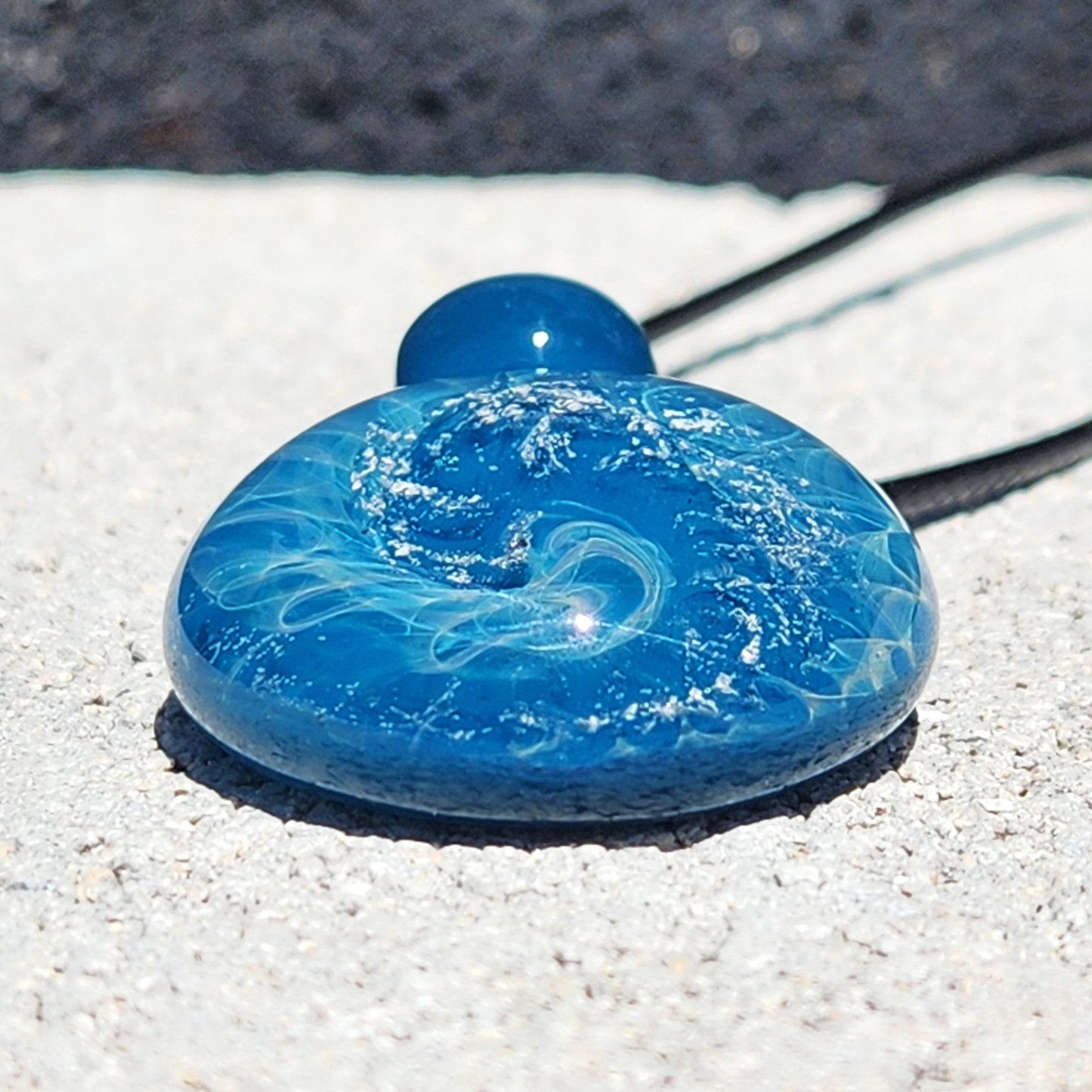 Close-up of handmade glass pendant with deep blue ocean wave patterns and silver flecks, resting on a light stone surface.