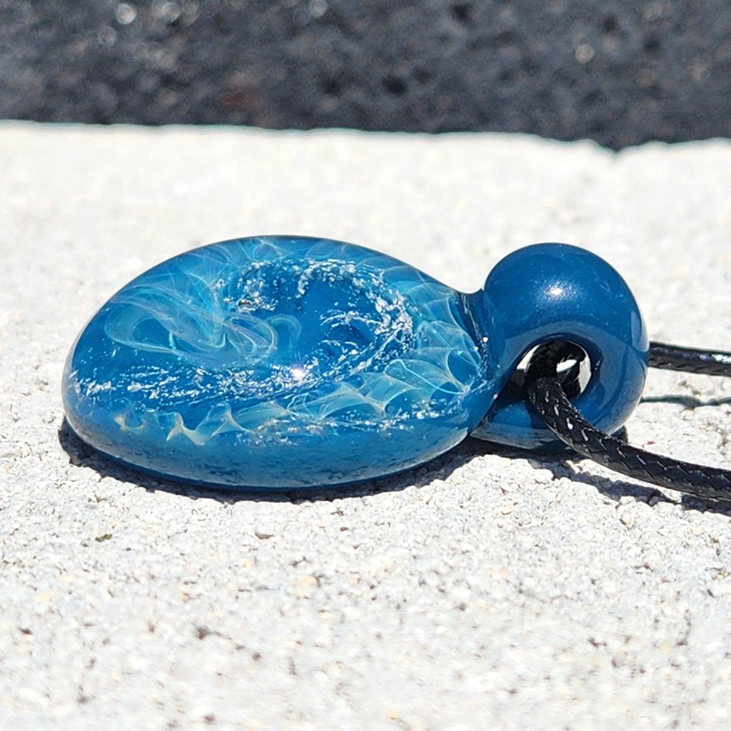 Angled view of blue glass pendant with swirling wave design and silver accents, resting on a sandy surface.