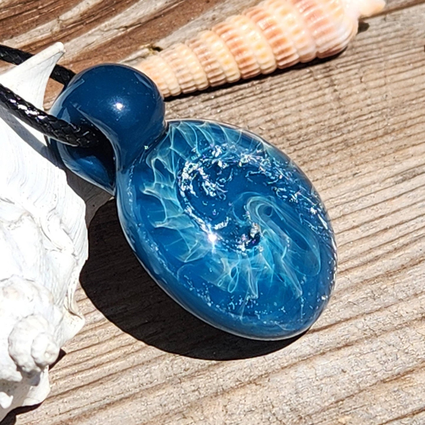 Ocean-inspired borosilicate glass pendant featuring blue swirls and silver accents, shown on a rustic wooden surface with seashells.