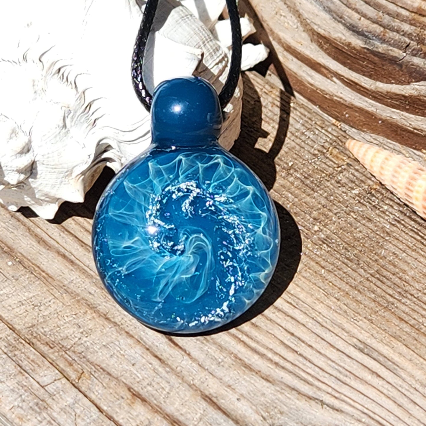Handmade borosilicate glass pendant with ocean wave motif and silver details, placed on wood with seashell decor.