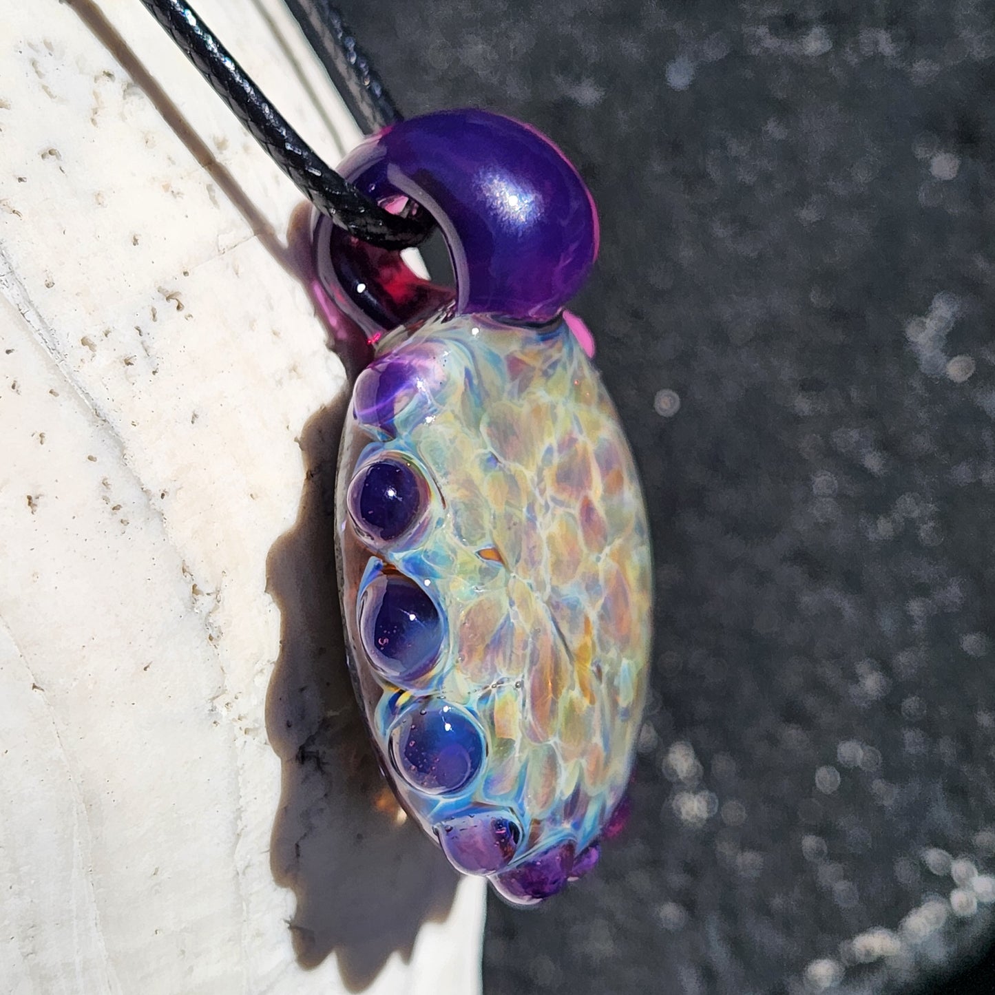 Angled view of handmade glass pendant with vibrant shimmer and purple accents, hanging on black cord. Funky borosilicate necklace design.