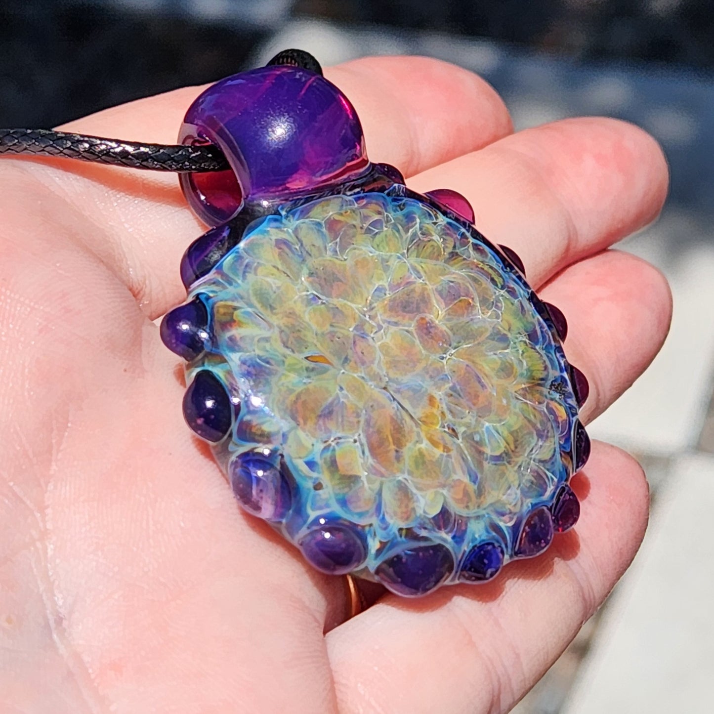 Close-up of large handmade glass pendant with multicolor shimmer and textured purple edge, shown in hand for scale. Vibrant borosilicate necklace.