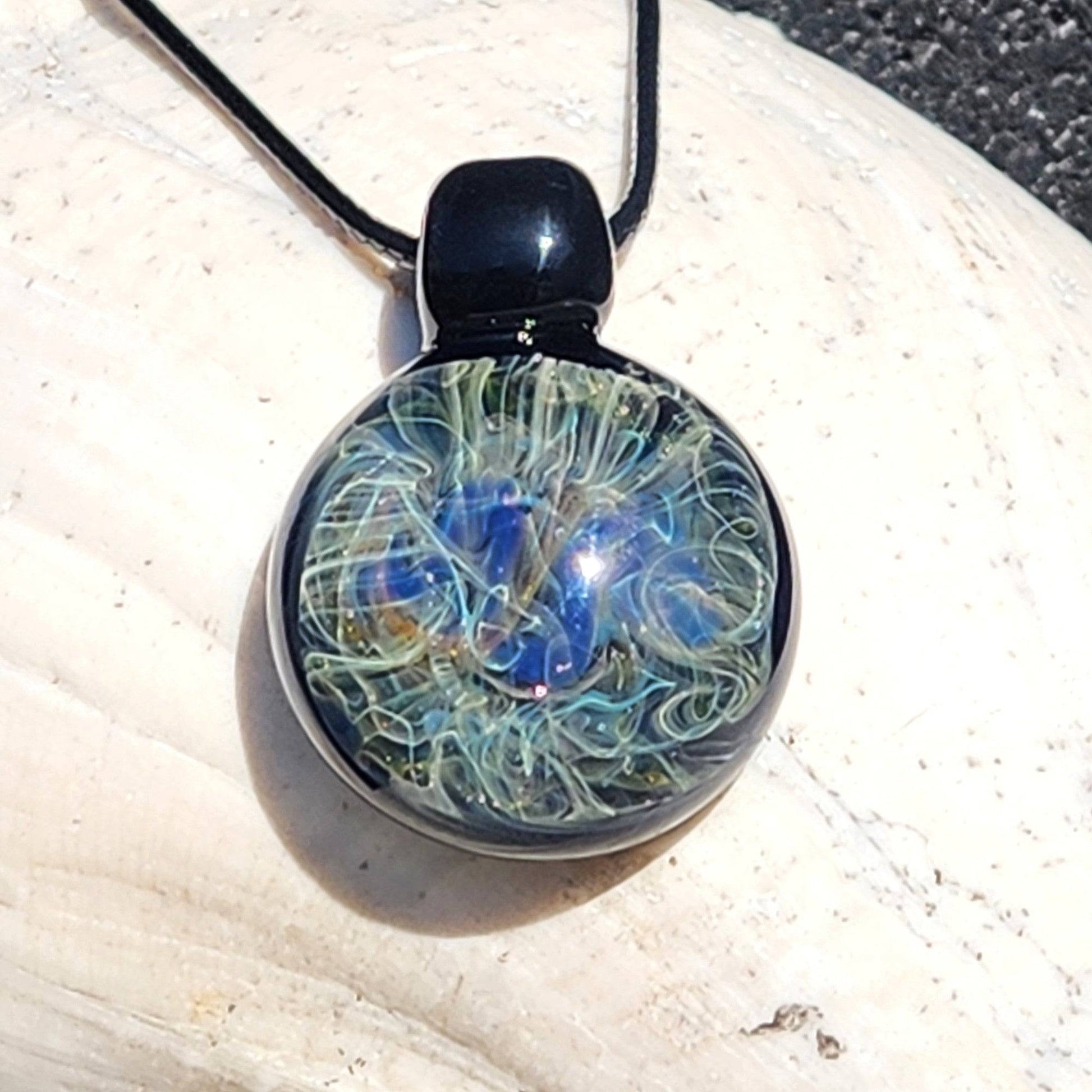 Round handmade glass pendant necklace with vibrant blue and gold swirls, suspended on black cord. Unique heady glass jewelry gift.