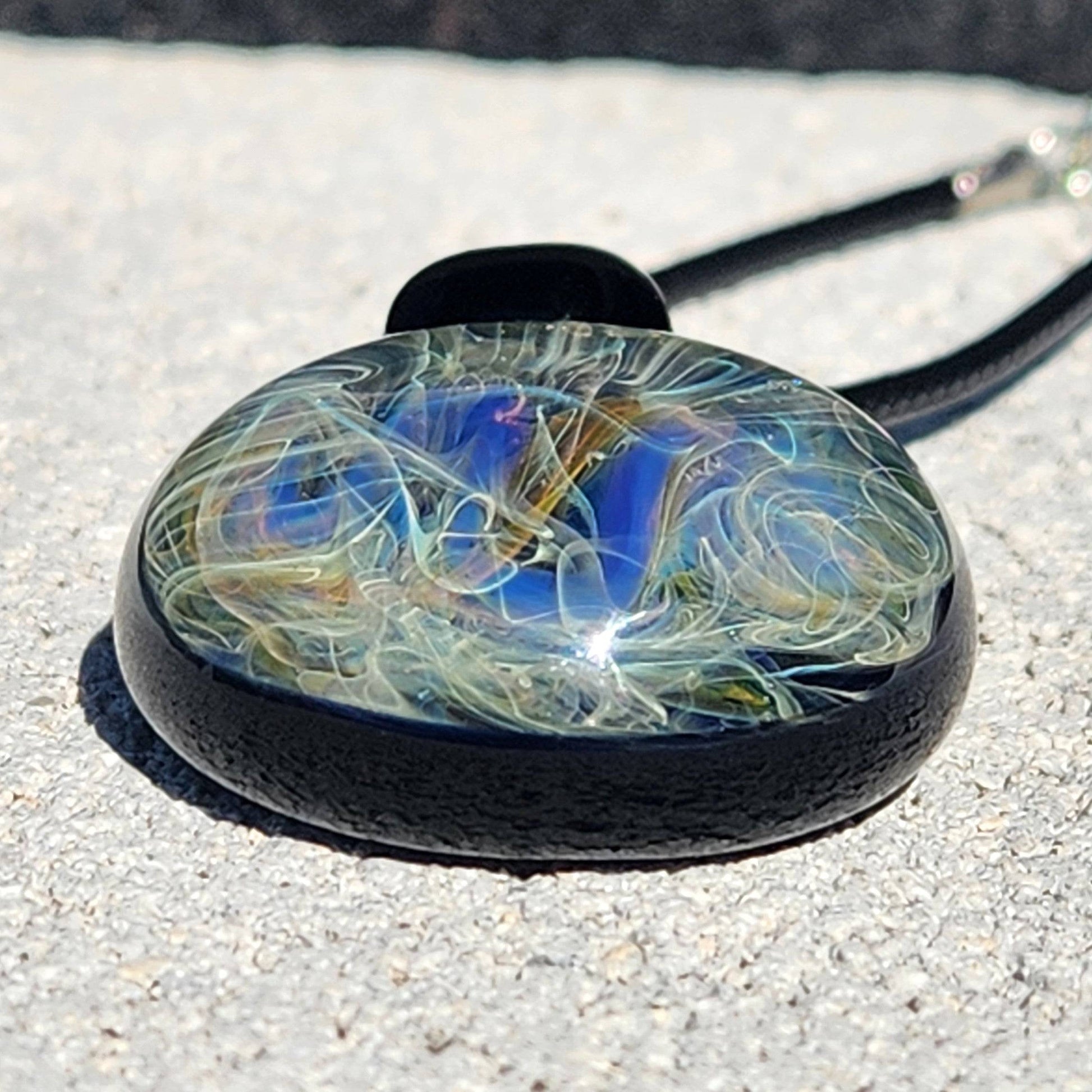Close-up of handmade blown glass pendant featuring intricate swirls of blue, gold, and silver on black base. Artistic jewelry for space lovers.