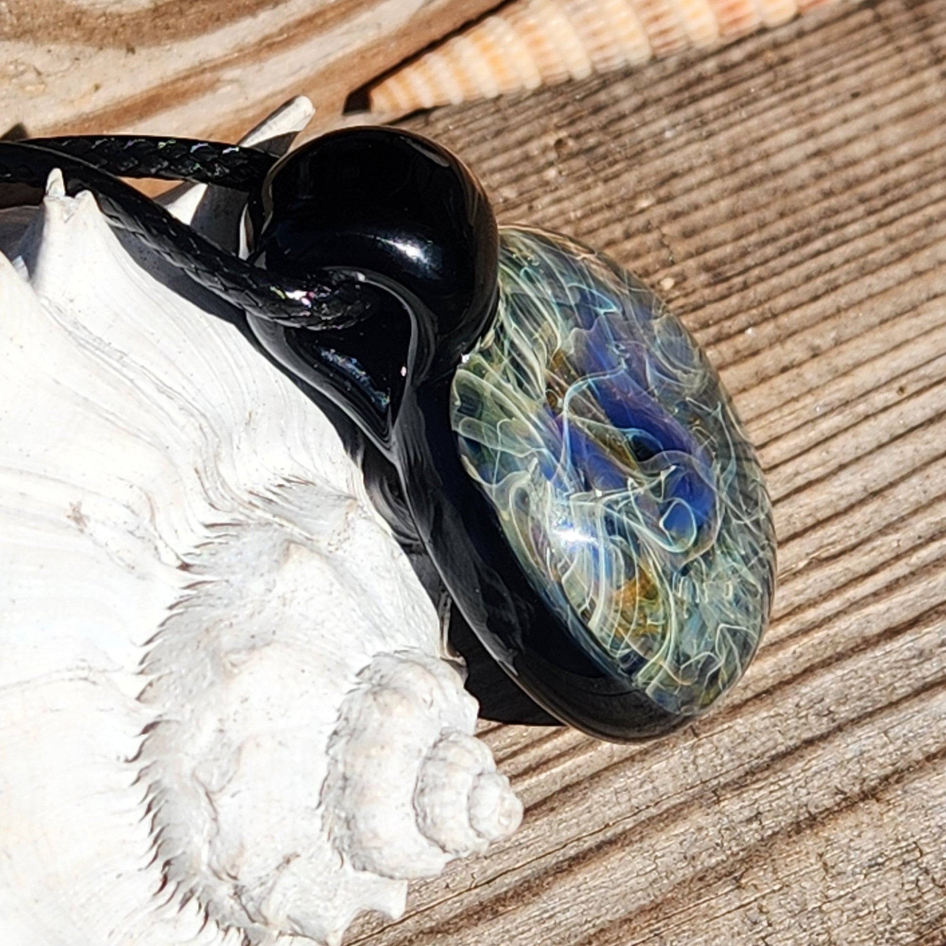 Handmade glass pendant necklace resting on seashell, showcasing intricate blue and gold swirls. Artistic heady glass jewelry piece.
