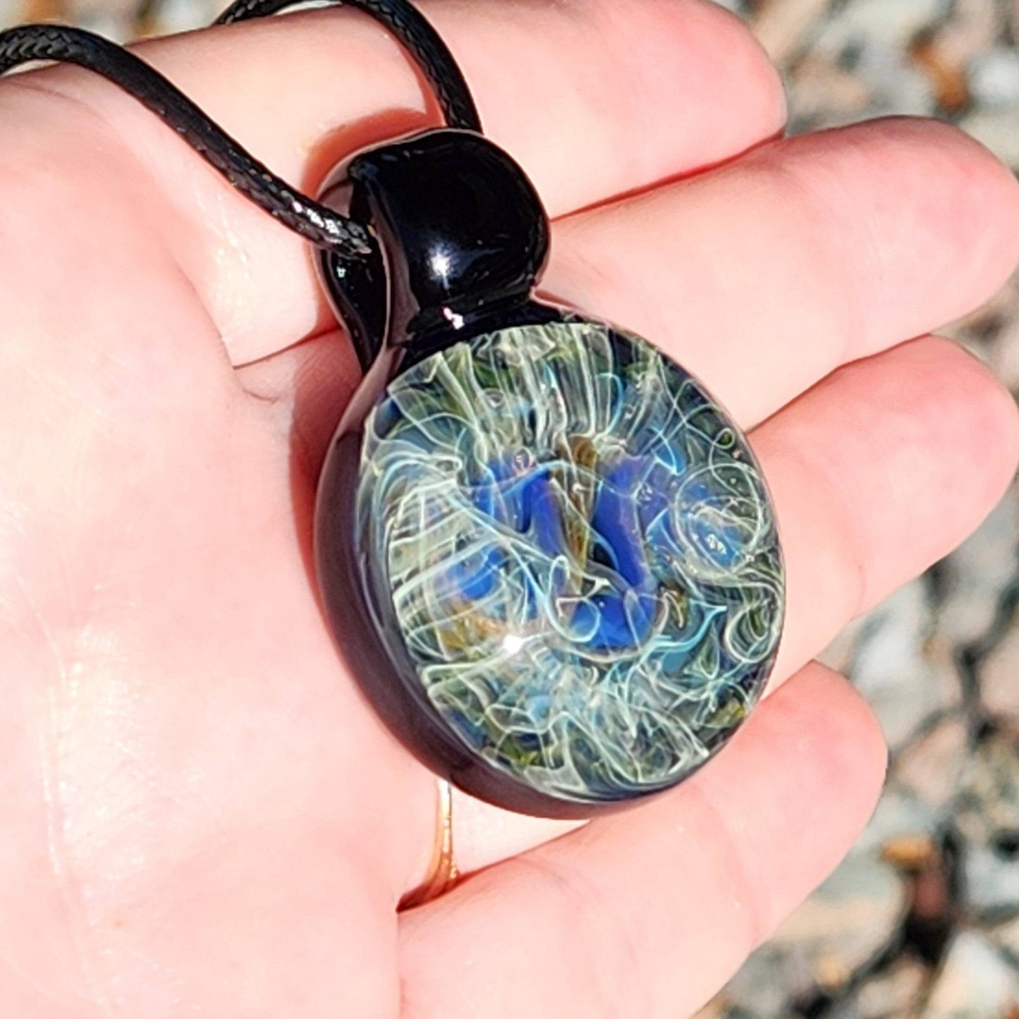 Close-up of pendant in hand, showing intricate handmade glass design with blue, gold, and silver swirls. Artistic necklace gift idea.