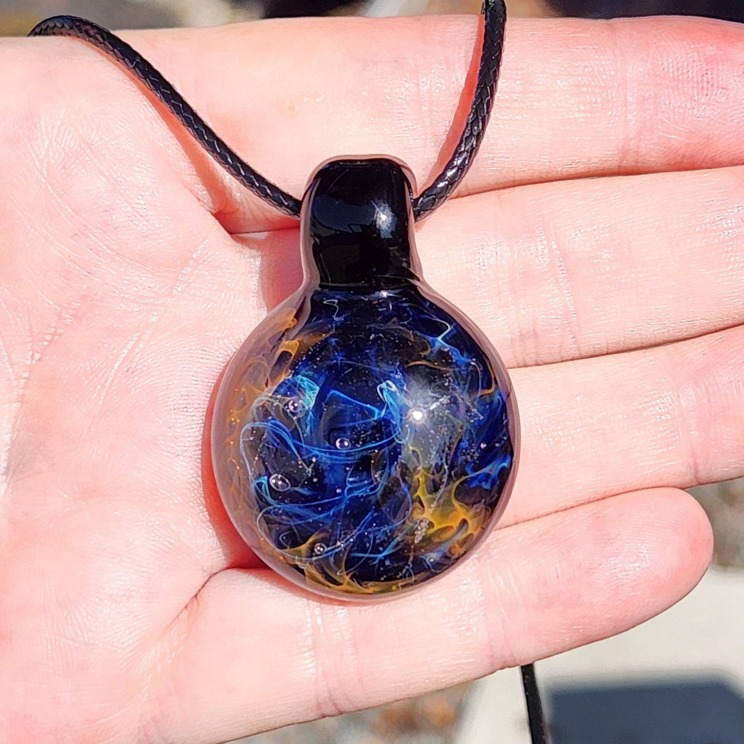 Hand holding a unique handmade blown glass pendant necklace with vibrant galaxy design in black, blue, and gold tones.