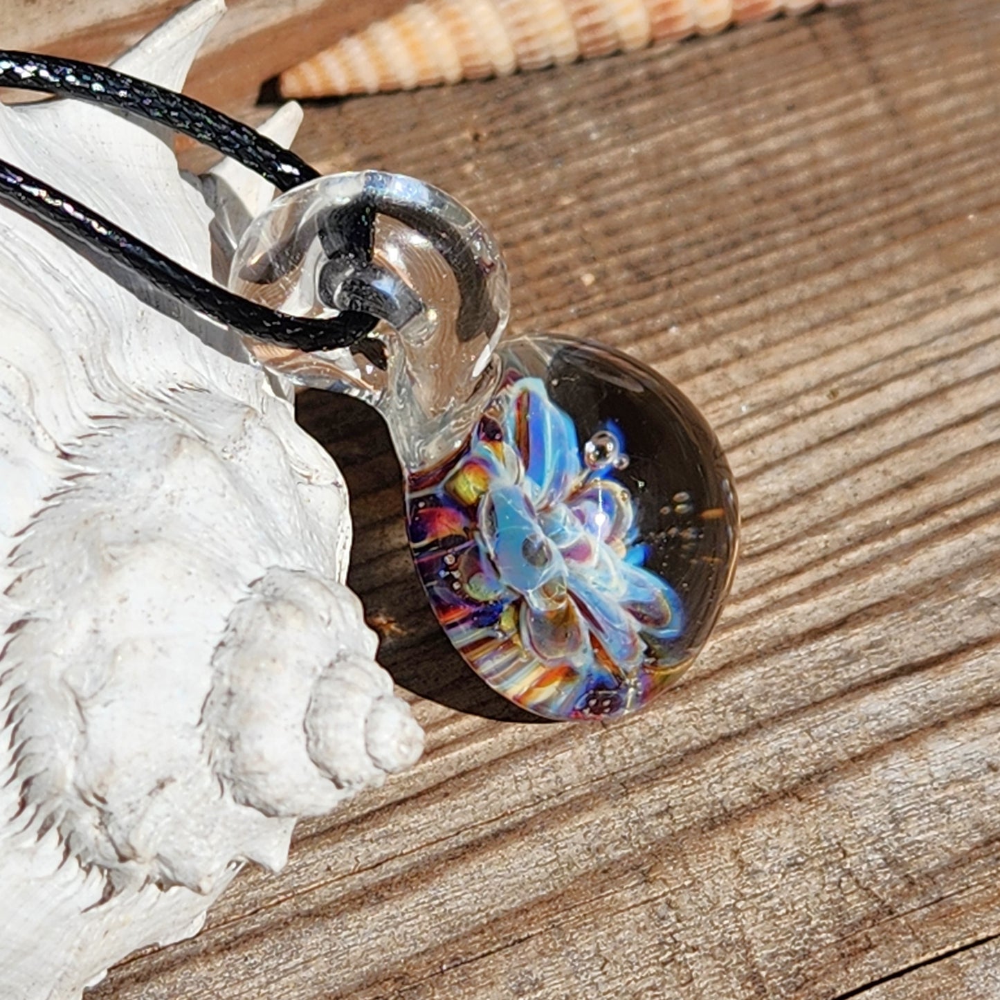 Angled view of UV Glow Glass Pendant on wood with shell, highlighting colorful flower. Unique handmade jewelry for fashion-forward wearers.