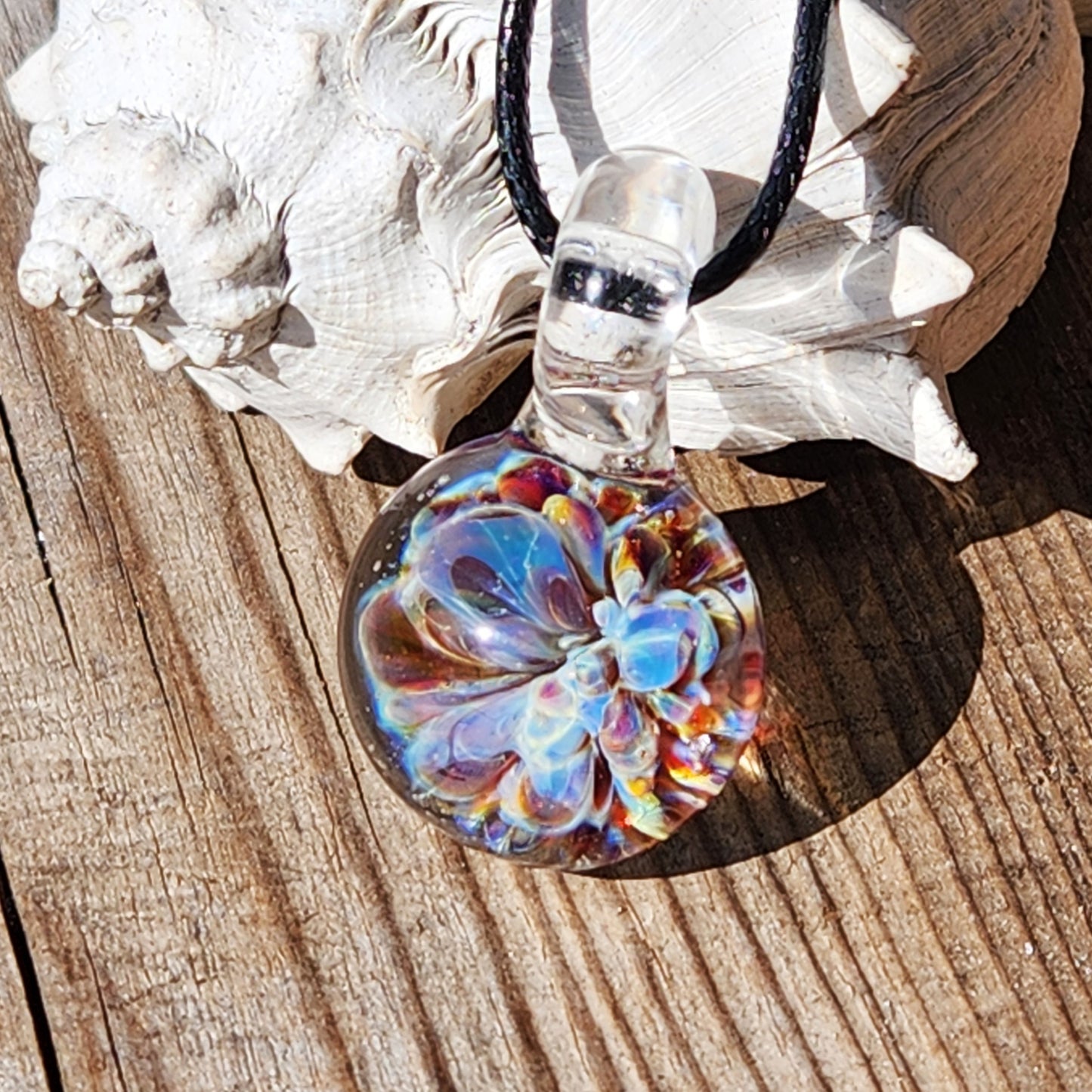UV Glow Glass Pendant resting on wood with shell background, showing vibrant flower inside. Handmade blown glass necklace for jewelry lovers.