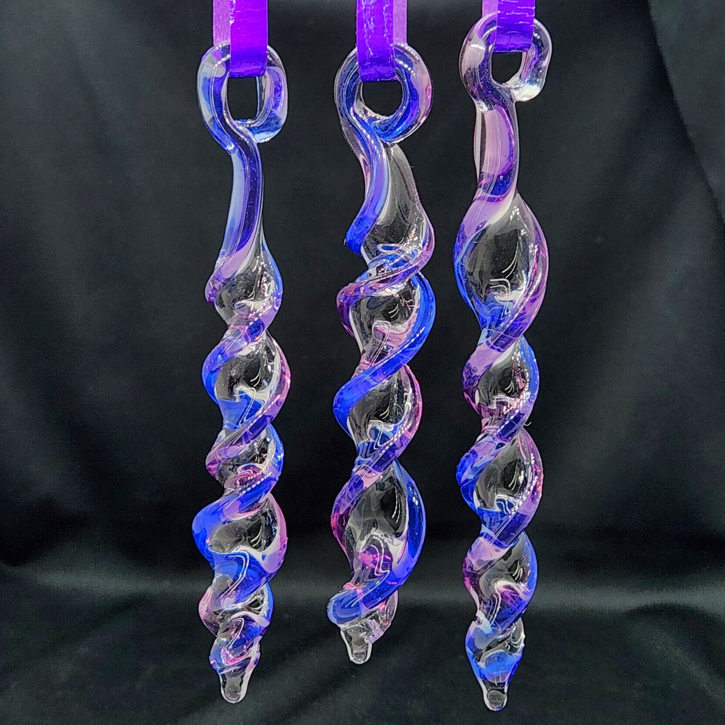 Three handmade glass Christmas ornaments in blue and purple with twisted icicle design, hanging against a dark background for contrast.