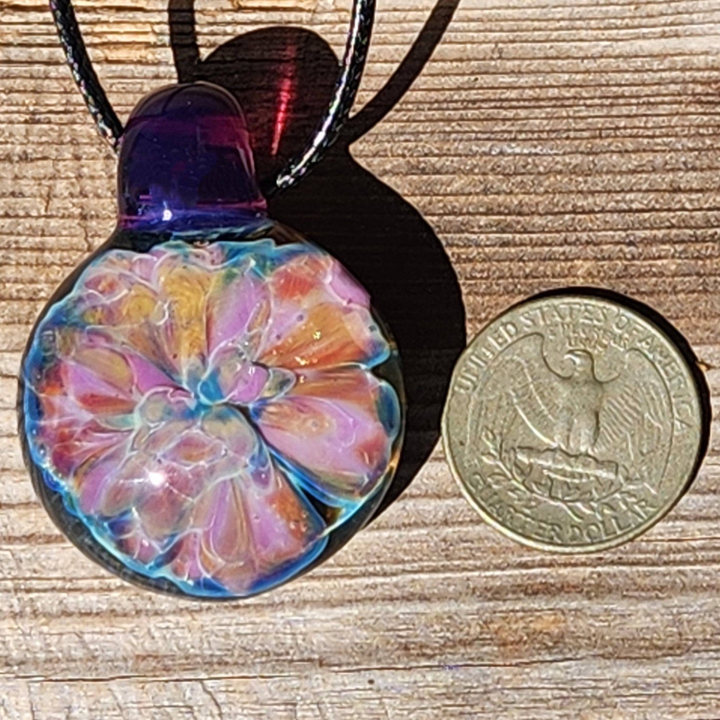 Handmade iridescent glass pendant necklace next to a U.S. quarter for size comparison, featuring a vibrant floral design and purple loop.