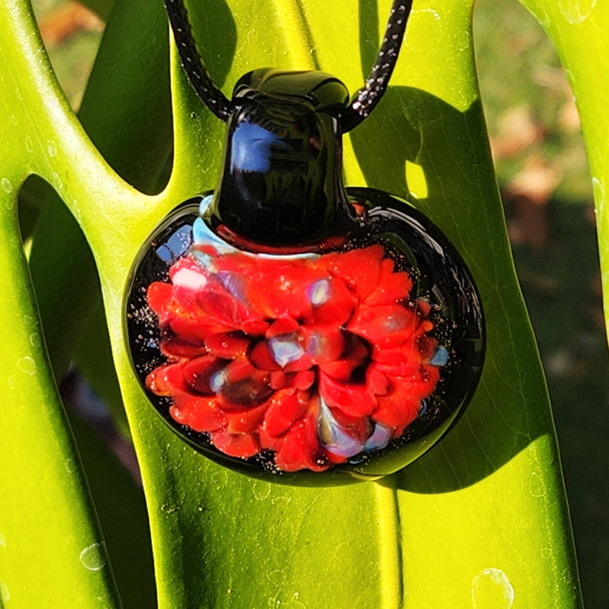 Red floral glass pendant necklace hanging on green leaf. Handmade borosilicate glass pendant with vibrant color and artistic craftsmanship.