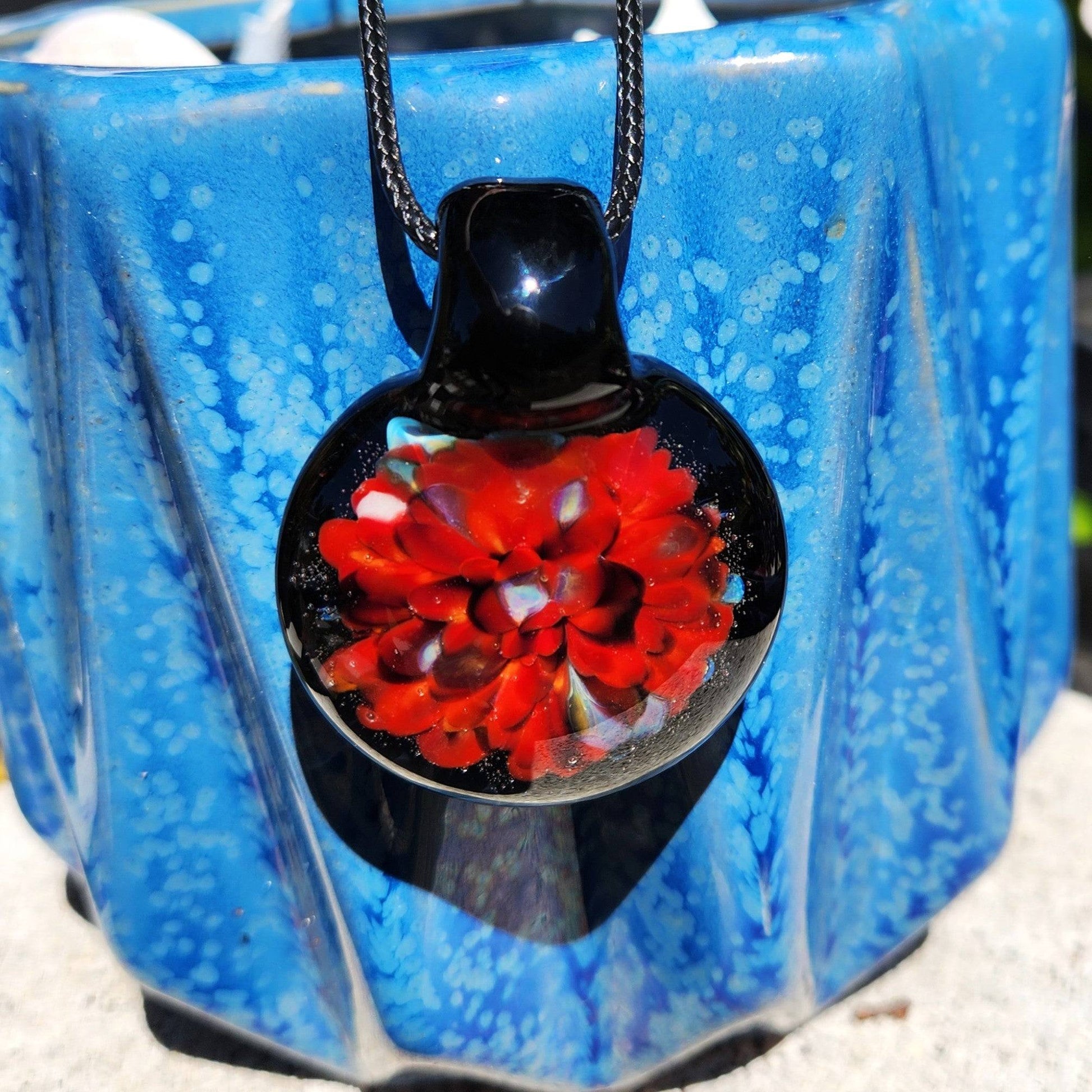 Radiant red glass pendant necklace displayed against a blue ceramic background. Handmade blown glass pendant with intricate floral detail.