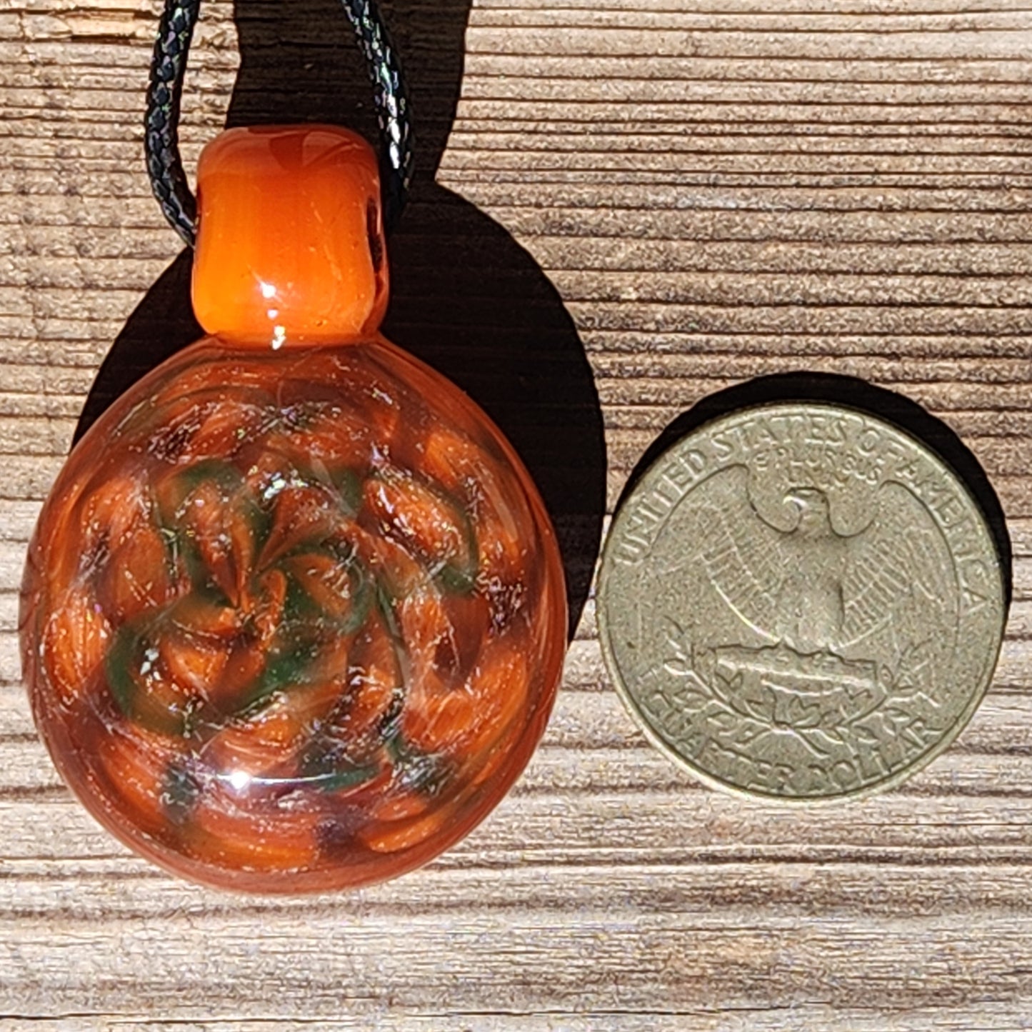 Size comparison of handmade orange swirl glass pendant necklace next to a U.S. half dollar coin on a wooden surface.