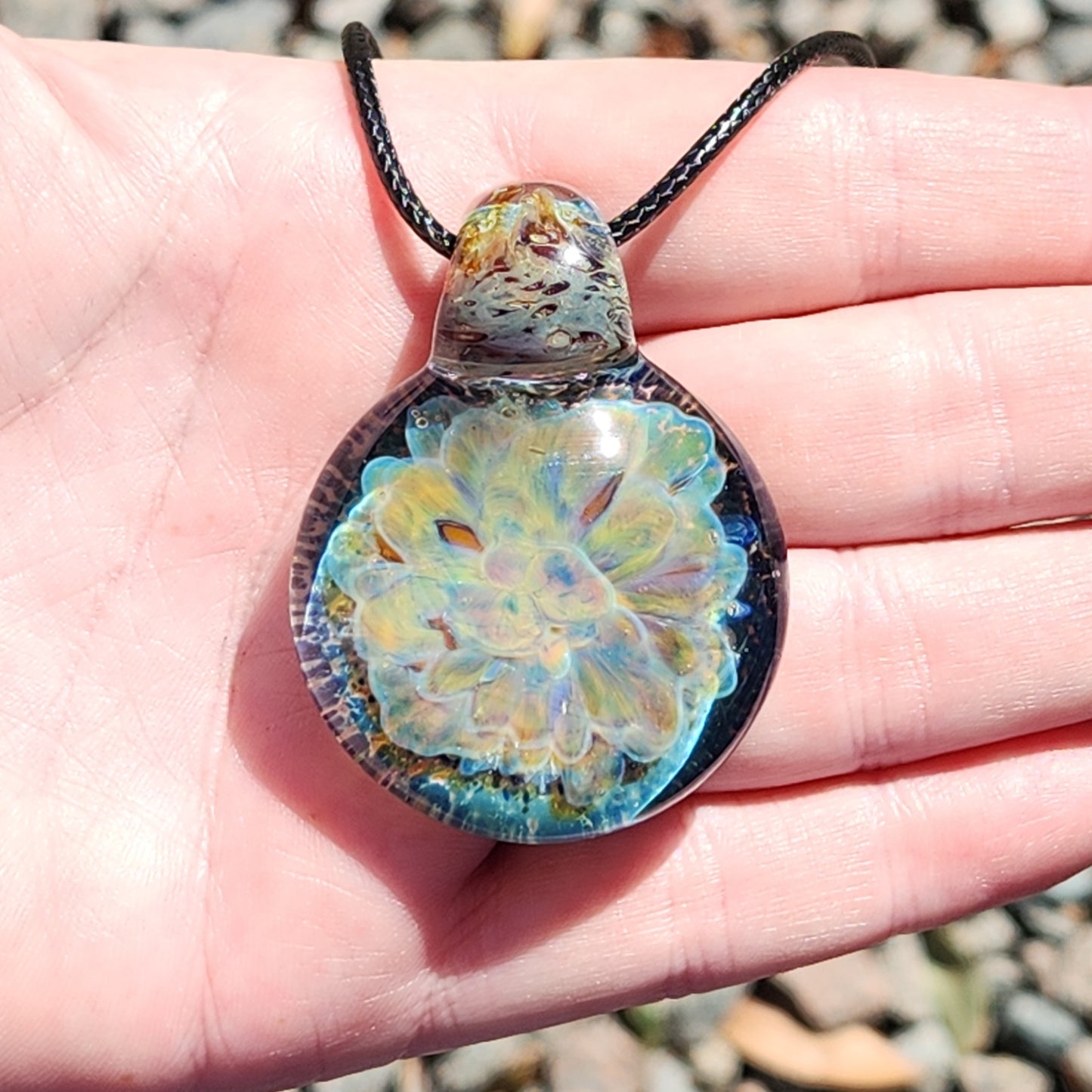 Close-up of the handmade glass pendant necklace in hand, showcasing the intricate yellow and blue floral pattern under sunlight.