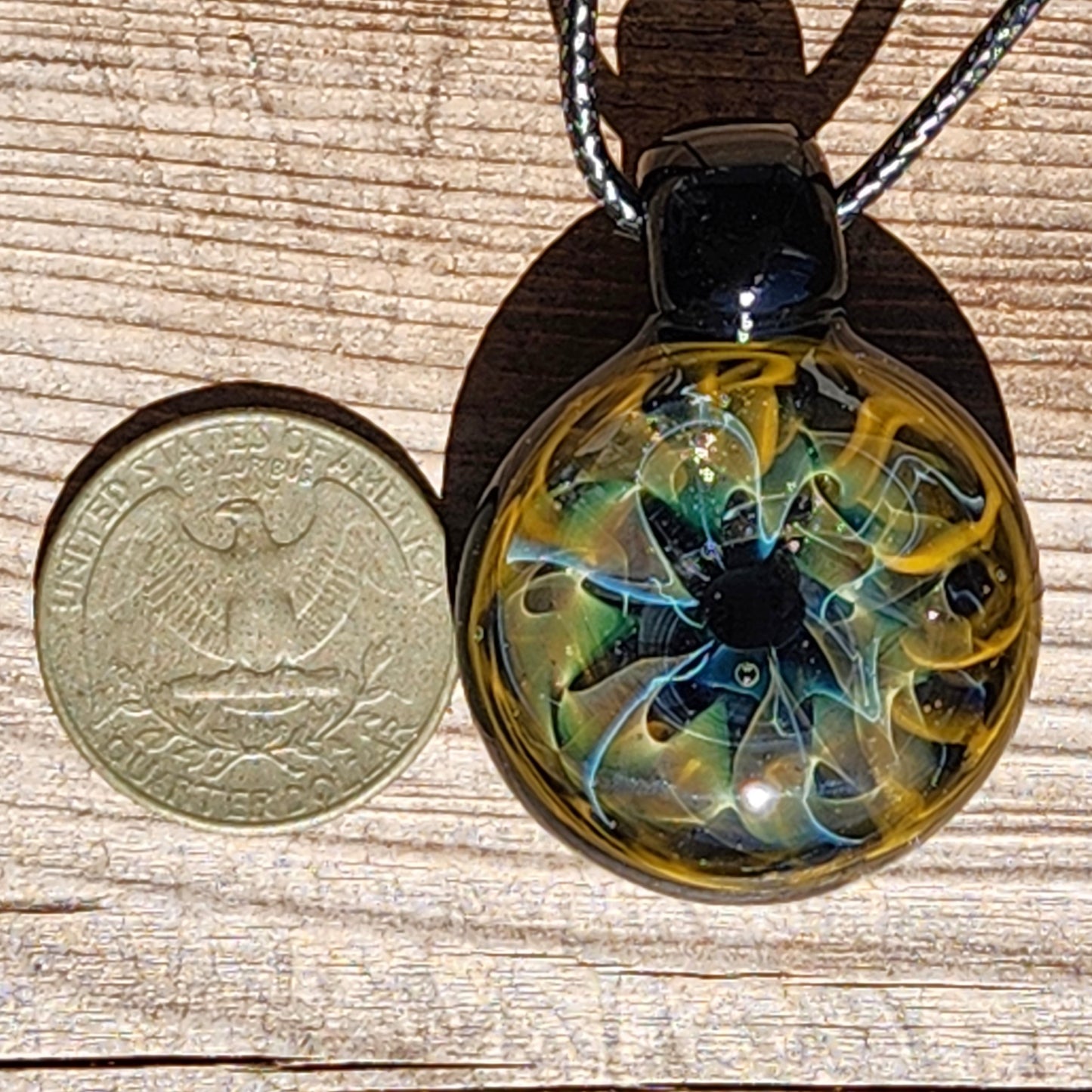 Unique handmade blown glass pendant necklace with swirling galaxy design in black, blue, and gold, shown next to a U.S. quarter for size reference.