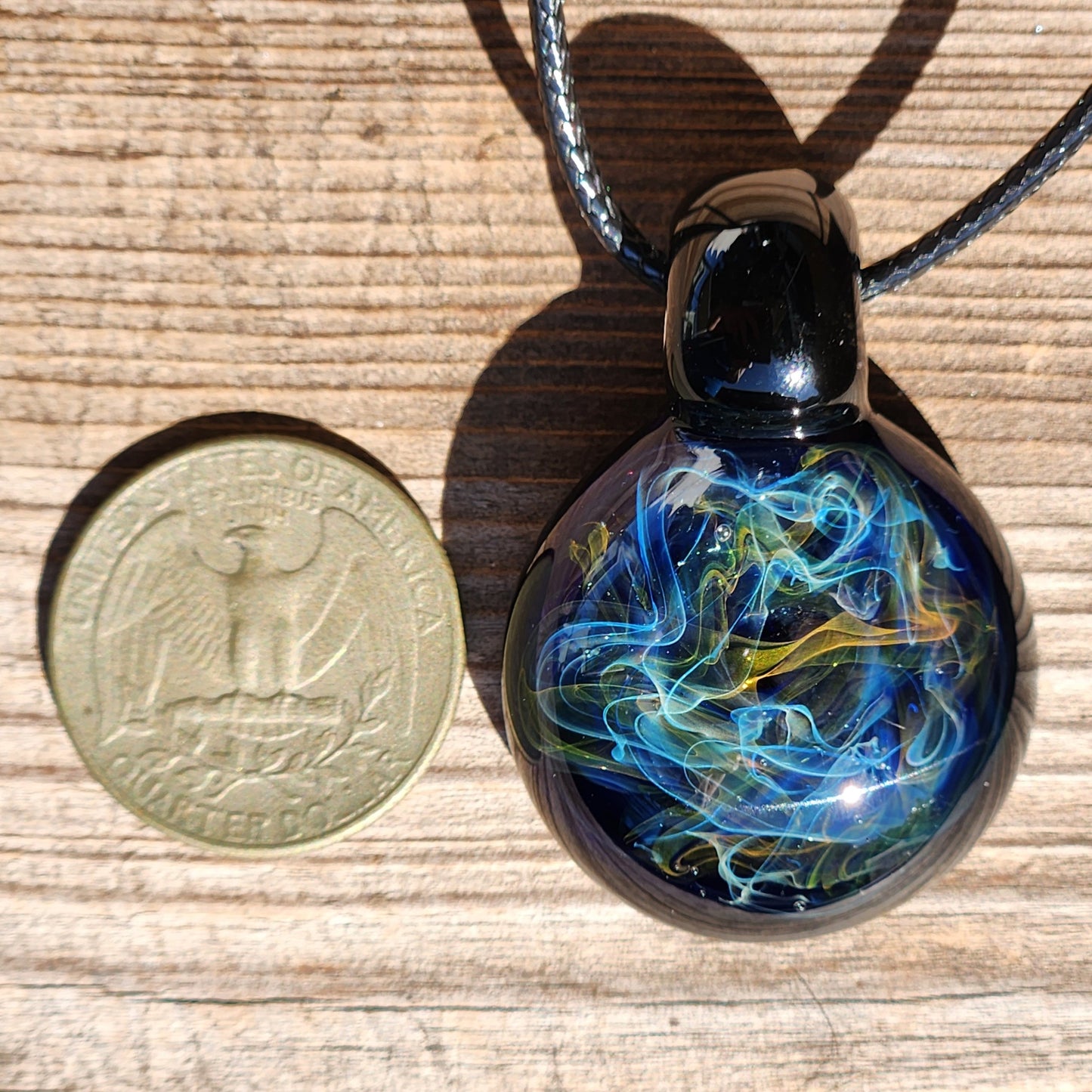 Side view of a black glass pendant with colorful swirling patterns, shadow cast on a wooden background, next to a coin for scale