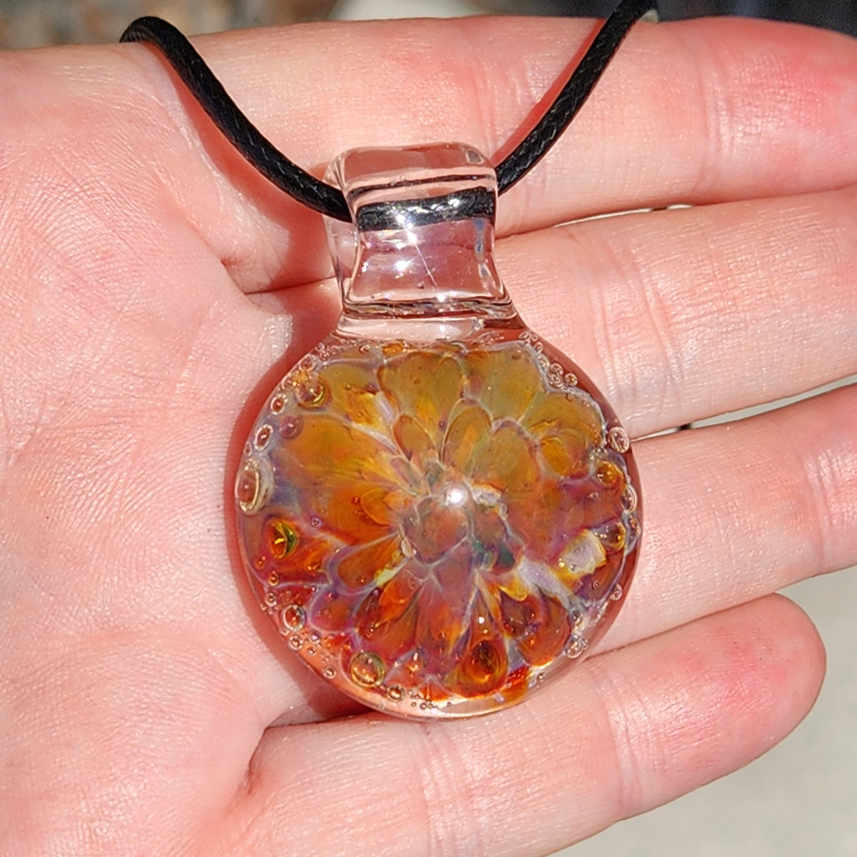 Detailed shot of the glass pendant necklace showing the vibrant sunset colors and the black cord, on a person's finger.