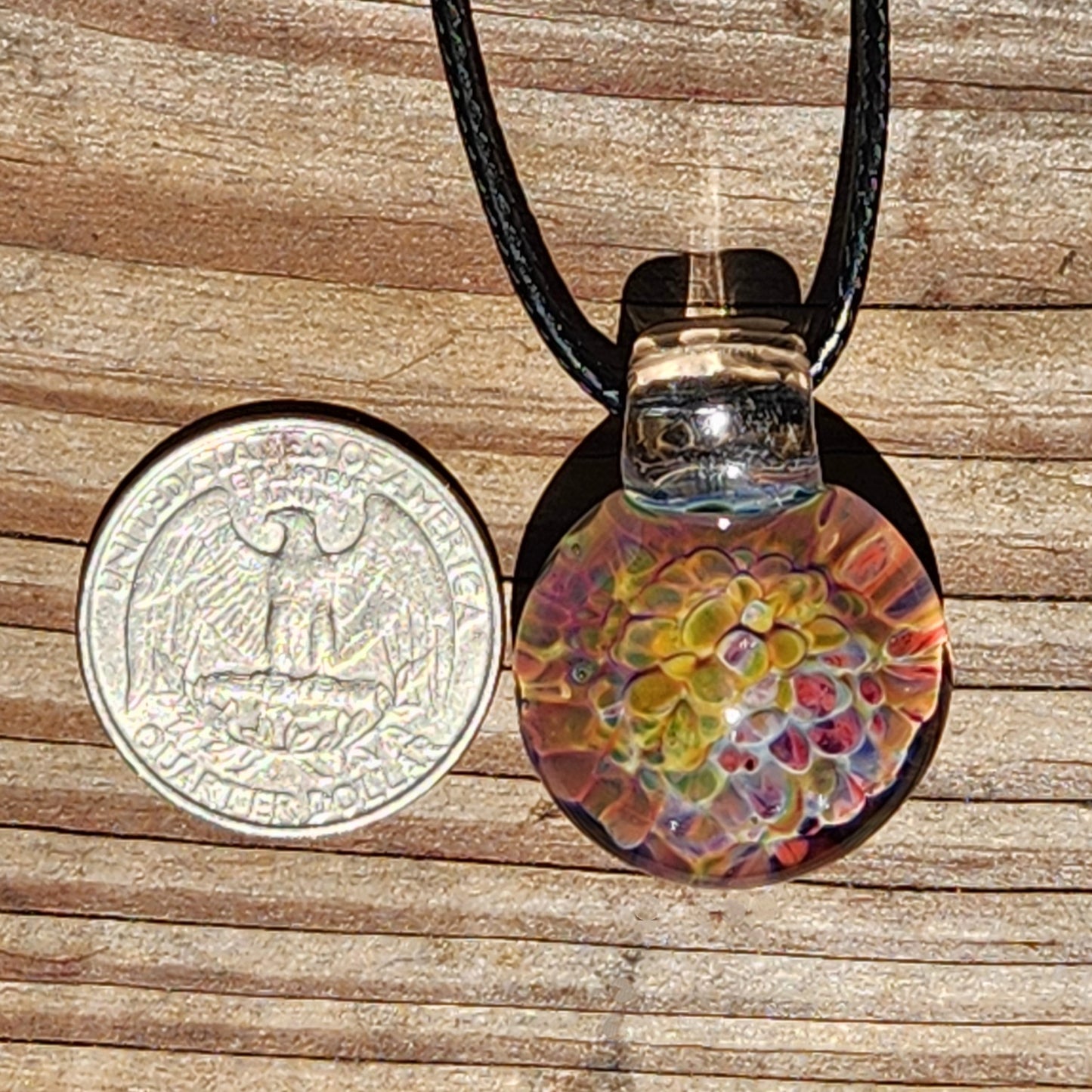 Colorful handmade glass pendant necklace with a rainbow swirl pattern, featuring vibrant hues of yellow, orange, pink, and purple, on a black cord, displayed on a wooden surface.