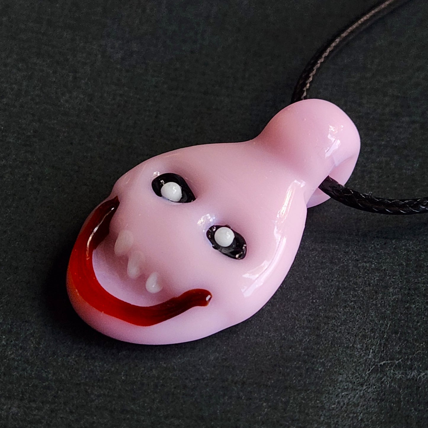 A glossy pink glass pendant shaped like a stylized face with white eyes, small teeth, and a wide red grin, hanging from a black cord on a dark textured background.