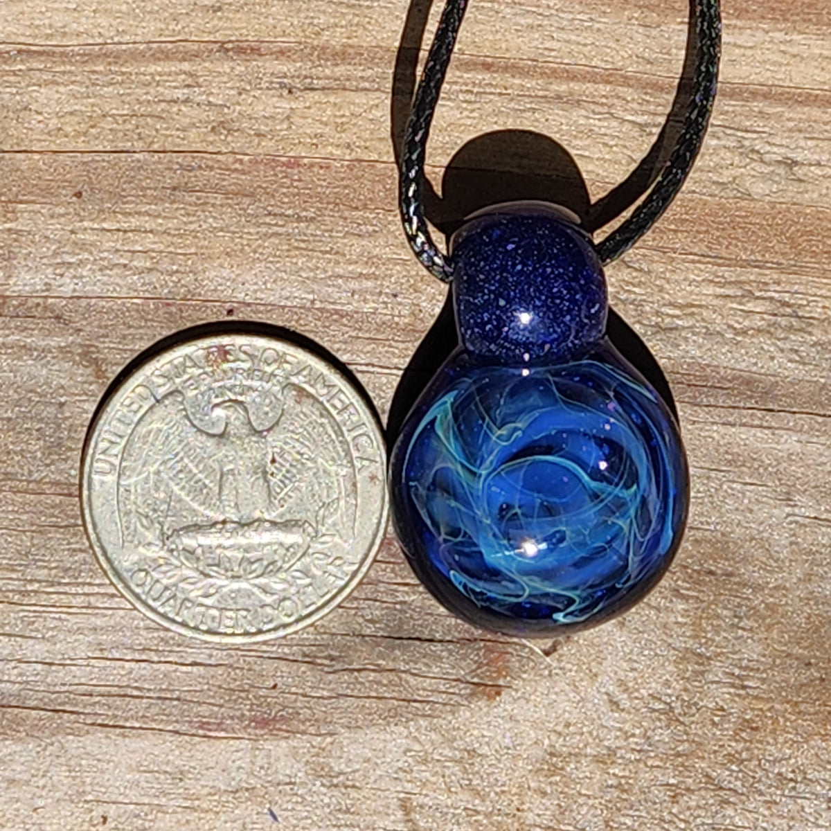 A blue glass pendant necklace with a black cord, shown next to a US quarter coin for scale on a wooden surface.