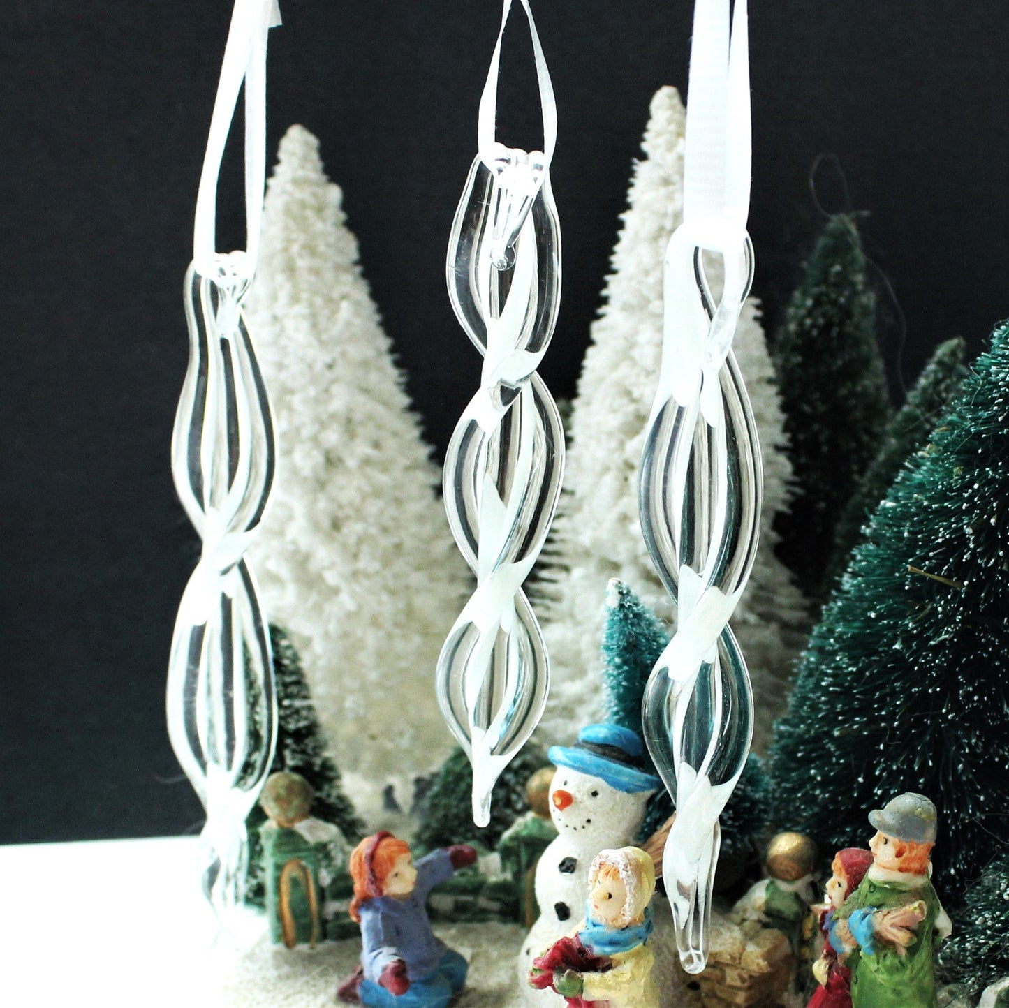Set of three clear spiral glass ornaments hanging among miniature snow-covered trees. Handmade Christmas keepsakes for festive home decor.