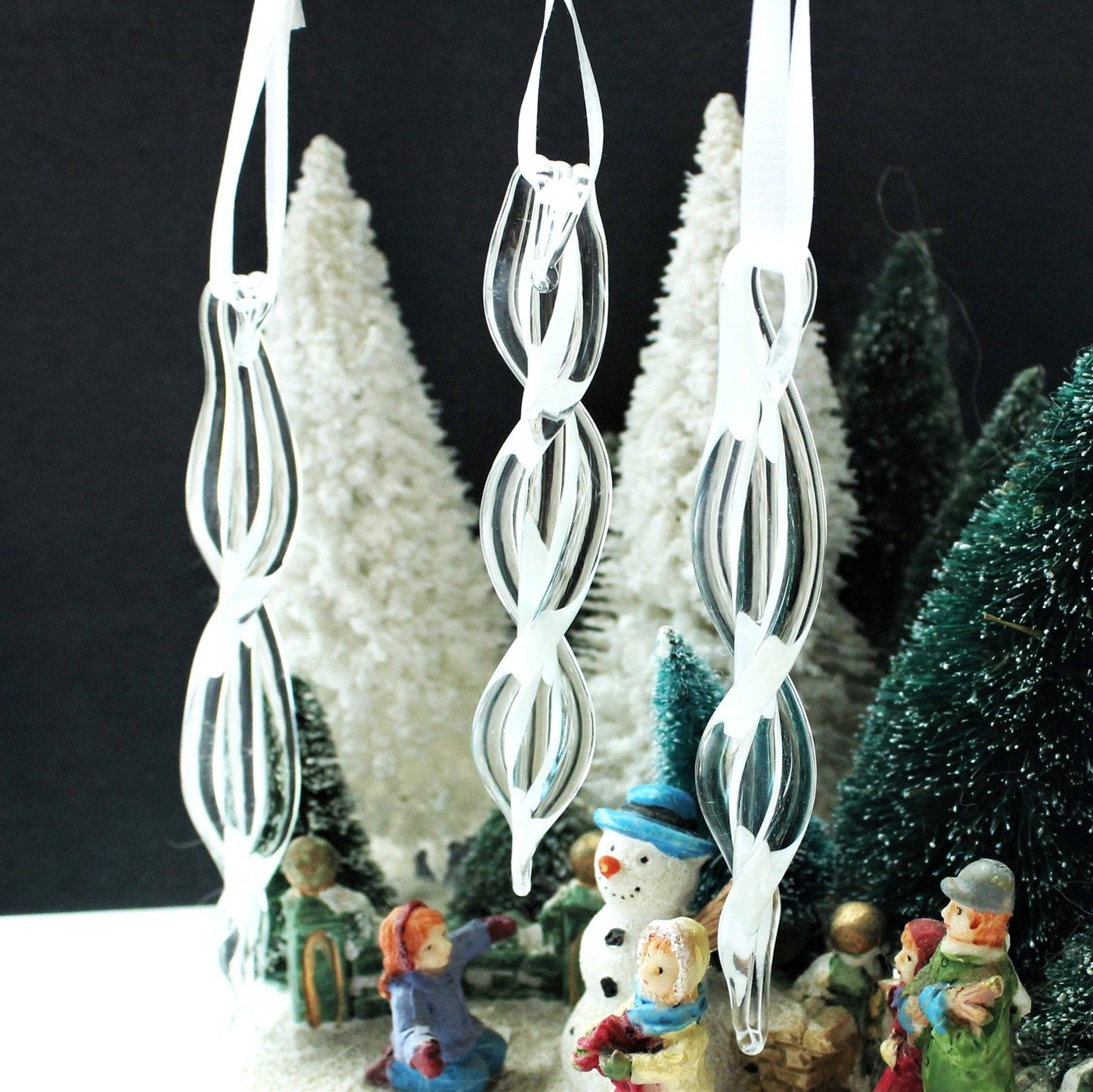 Set of three clear spiral glass ornaments hanging among miniature snow-covered trees. Handmade Christmas keepsakes for festive home decor.