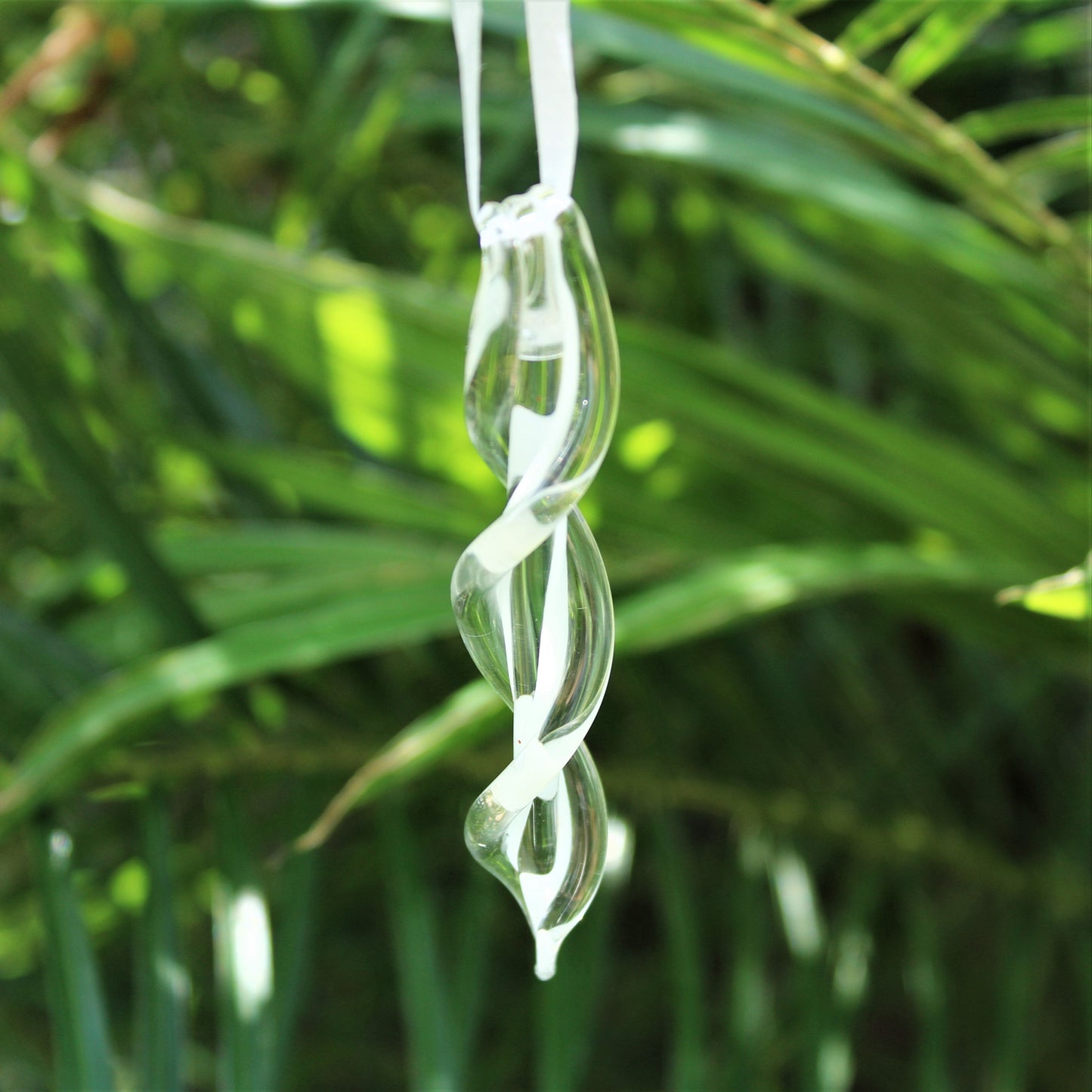 Clear handmade glass ornament with spiral design hanging in front of green foliage. Glow-in-the-dark Christmas keepsake for indoor or outdoor use.