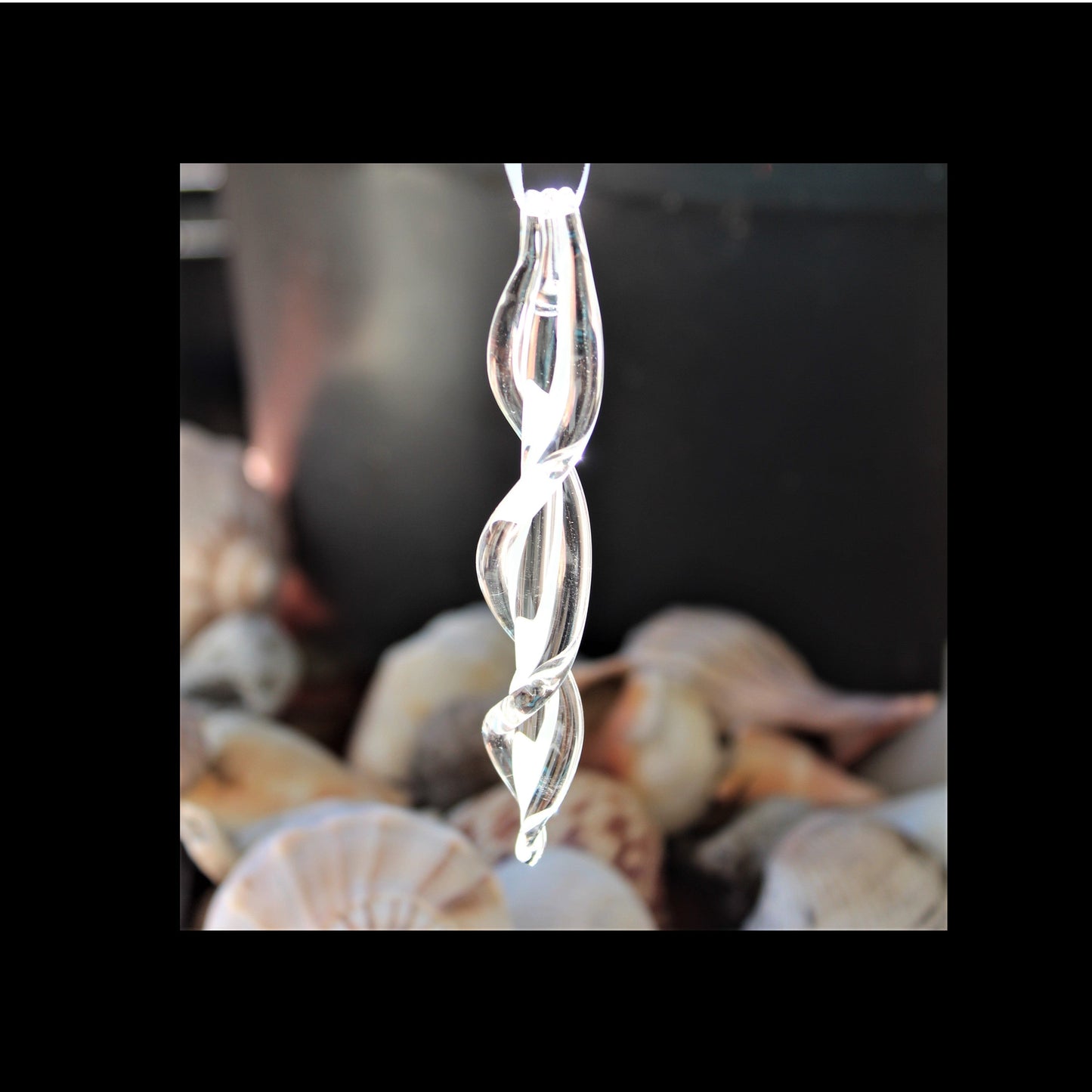 Clear handmade glass ornament with spiral twist design hanging in front of seashells. Ideal for Christmas or coastal-themed holiday decorations.